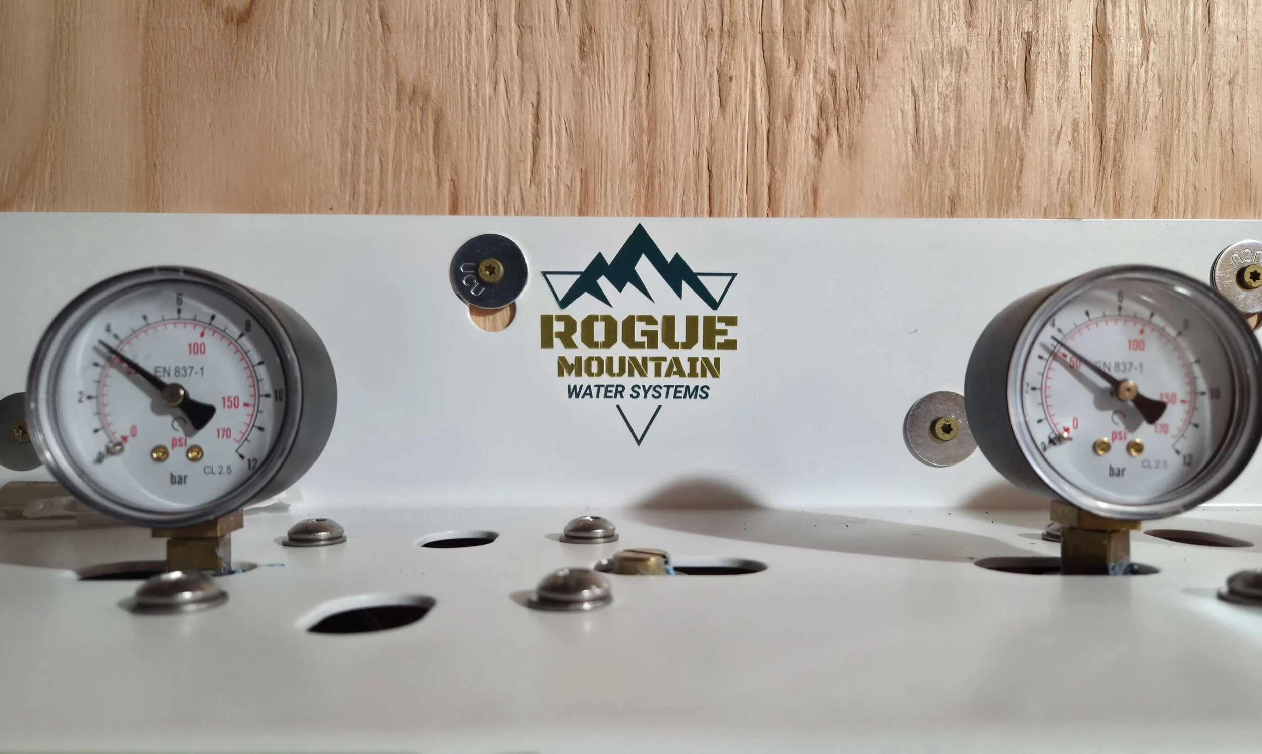 A white panel with two pressure gauges labeled in psi and bar, with the brand 'Rogue Mountain Water Systems' logo on the front, mounted on a wooden surface.