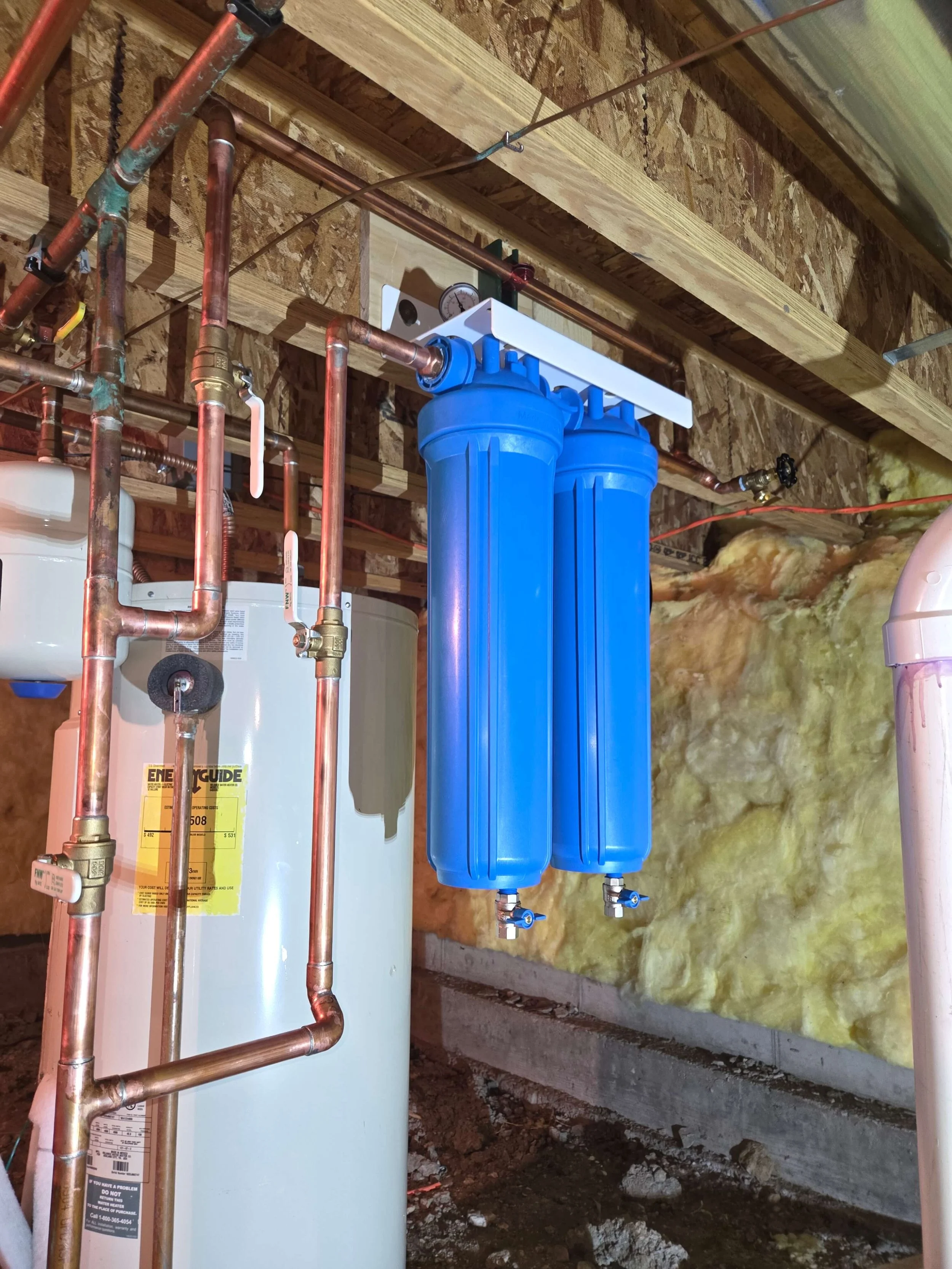 Water filtration system with blue filters, copper pipes, a pressure gauge, and insulation in a building's basement or utility area.