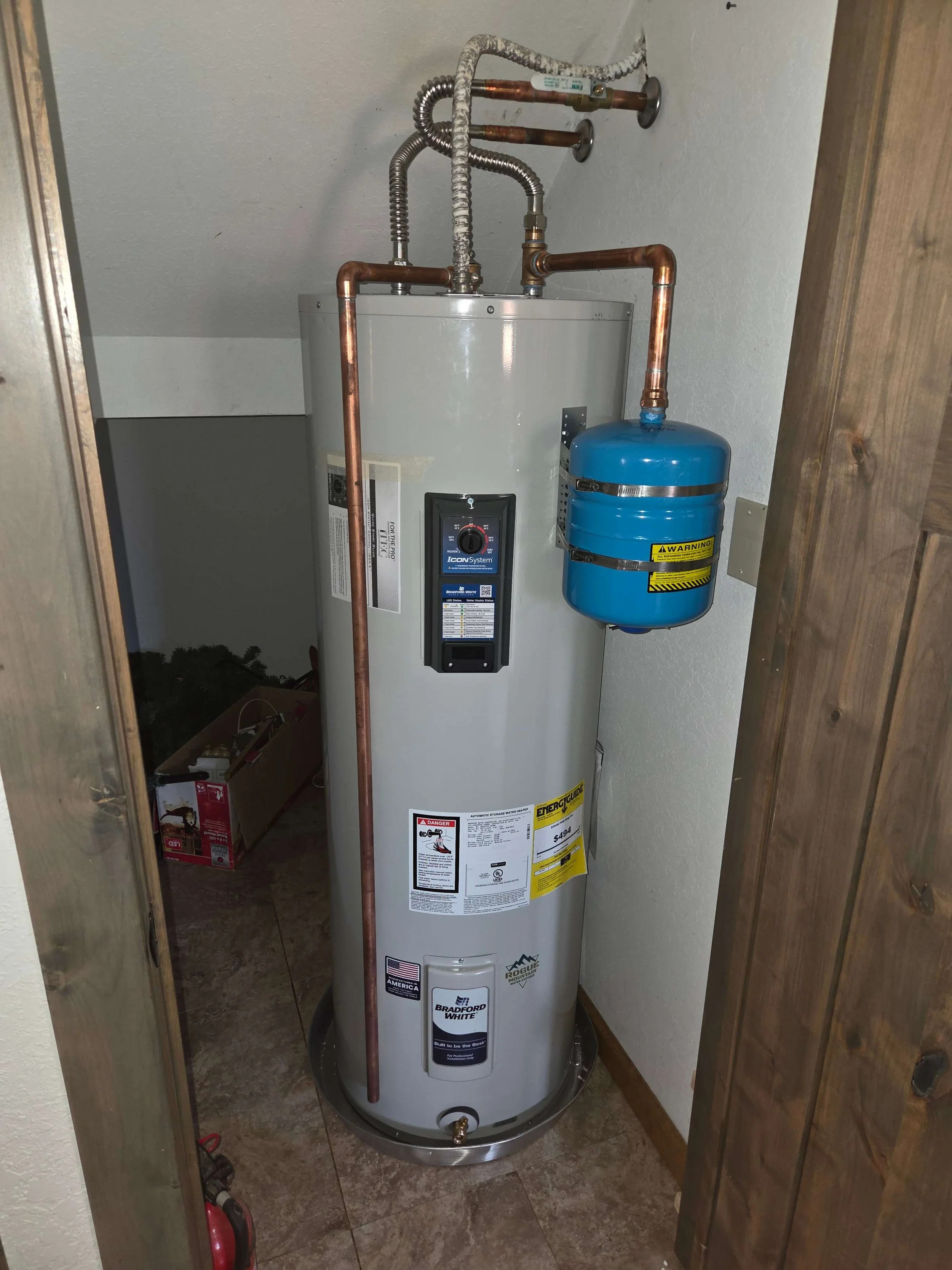 A water heater with copper pipes and a blue expansion tank installed in a closet with wood paneling on the sides.