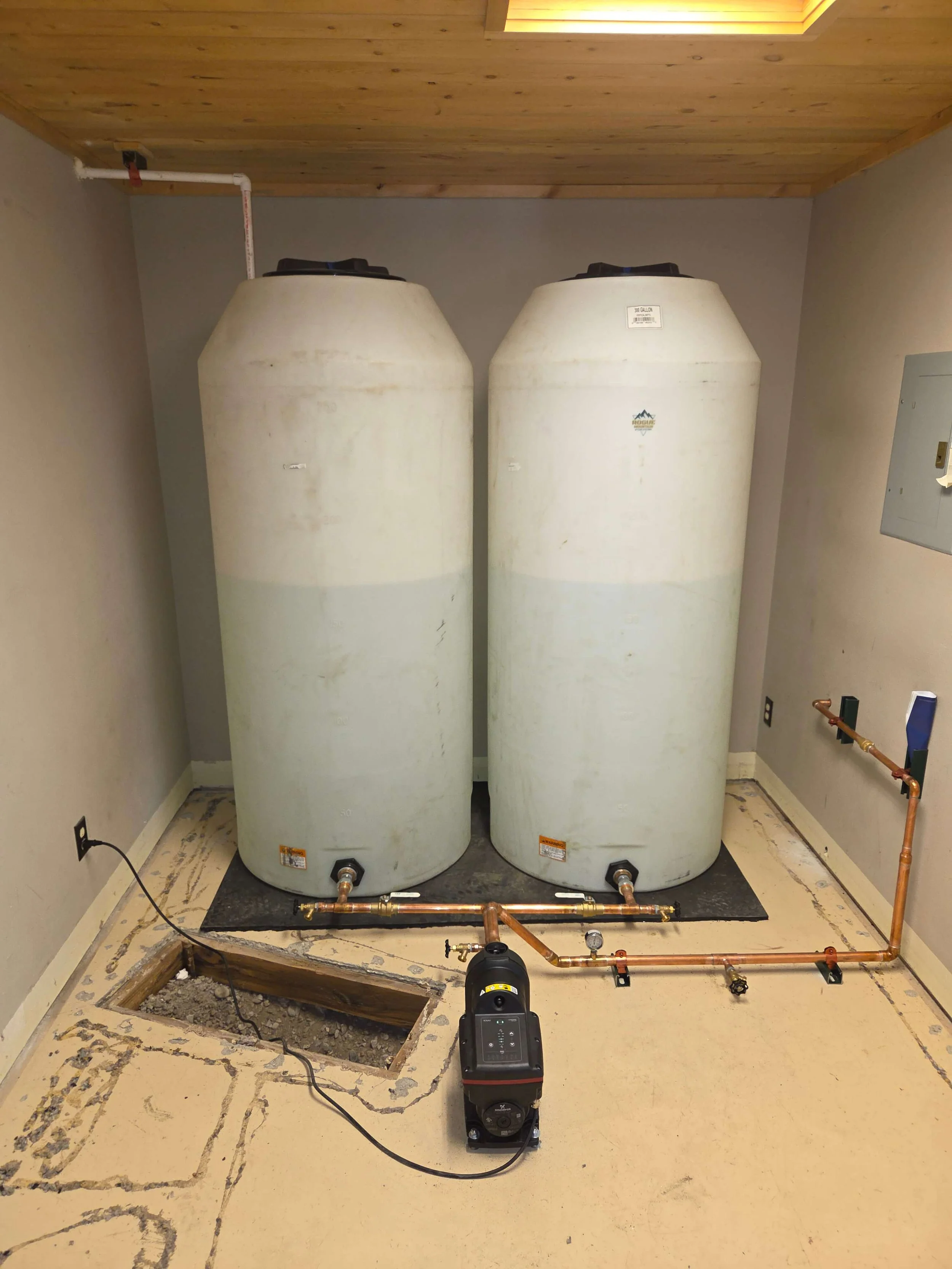 Two large white water tanks in a room, with copper piping and a black pump on the floor, and an access hole in the floor with exposed wiring and insulation around the opening.