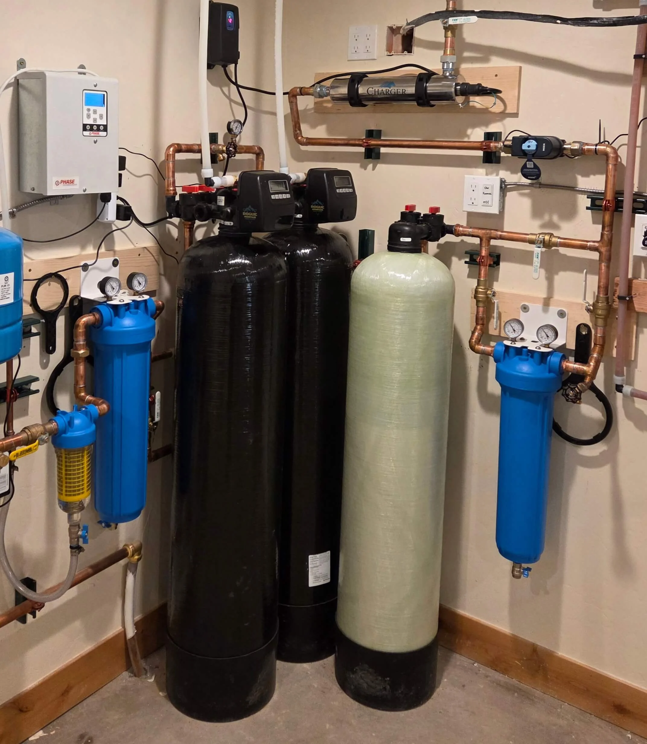 A water treatment system featuring three large vertical tanks, two black and one green, connected by copper pipes, with gauges, valves, and control units mounted on a beige wall in a utility room.