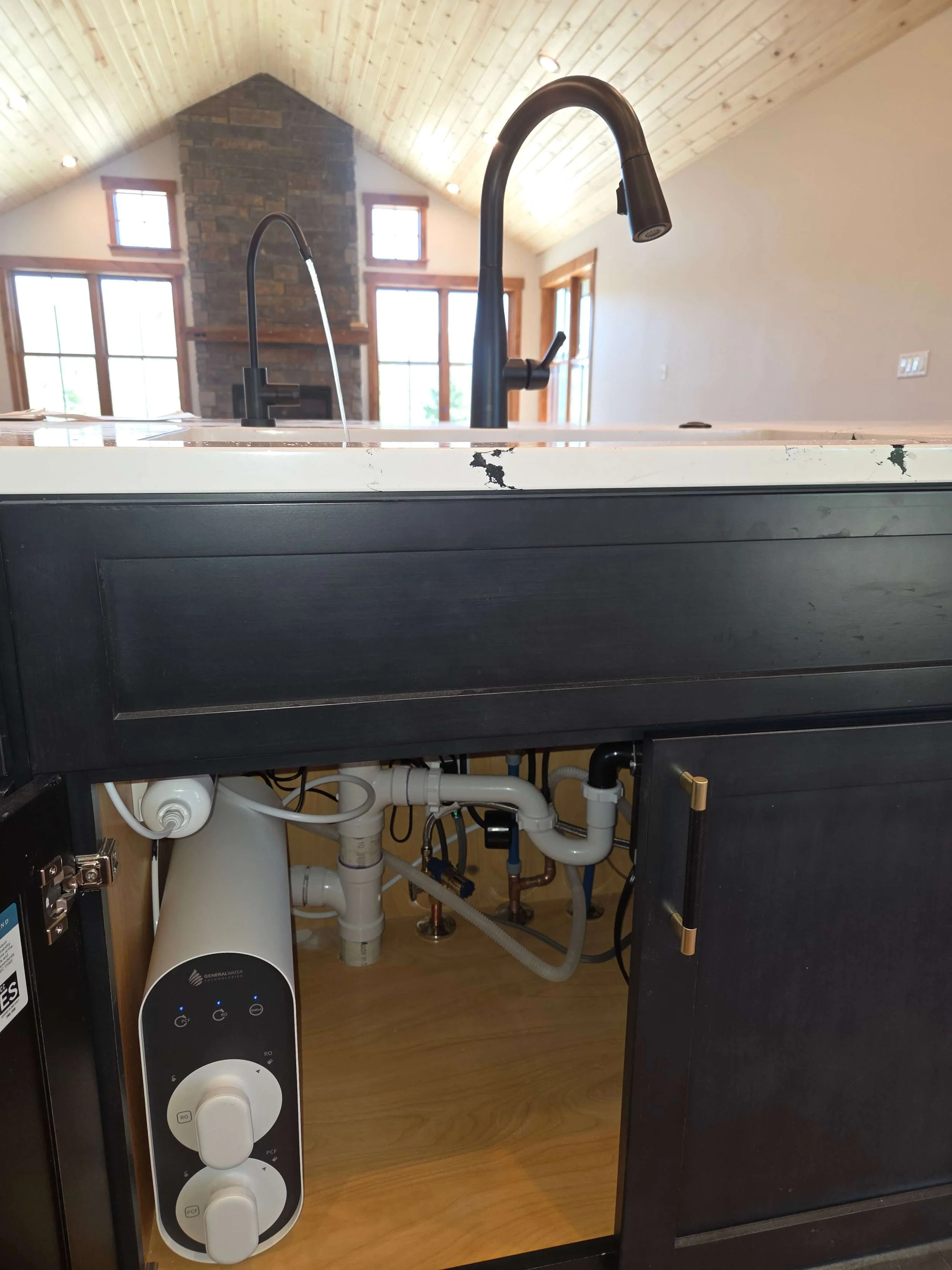 View of a kitchen sink with black faucets, showing plumbing and water filtration system underneath the sink in a room with vaulted ceiling and large windows.