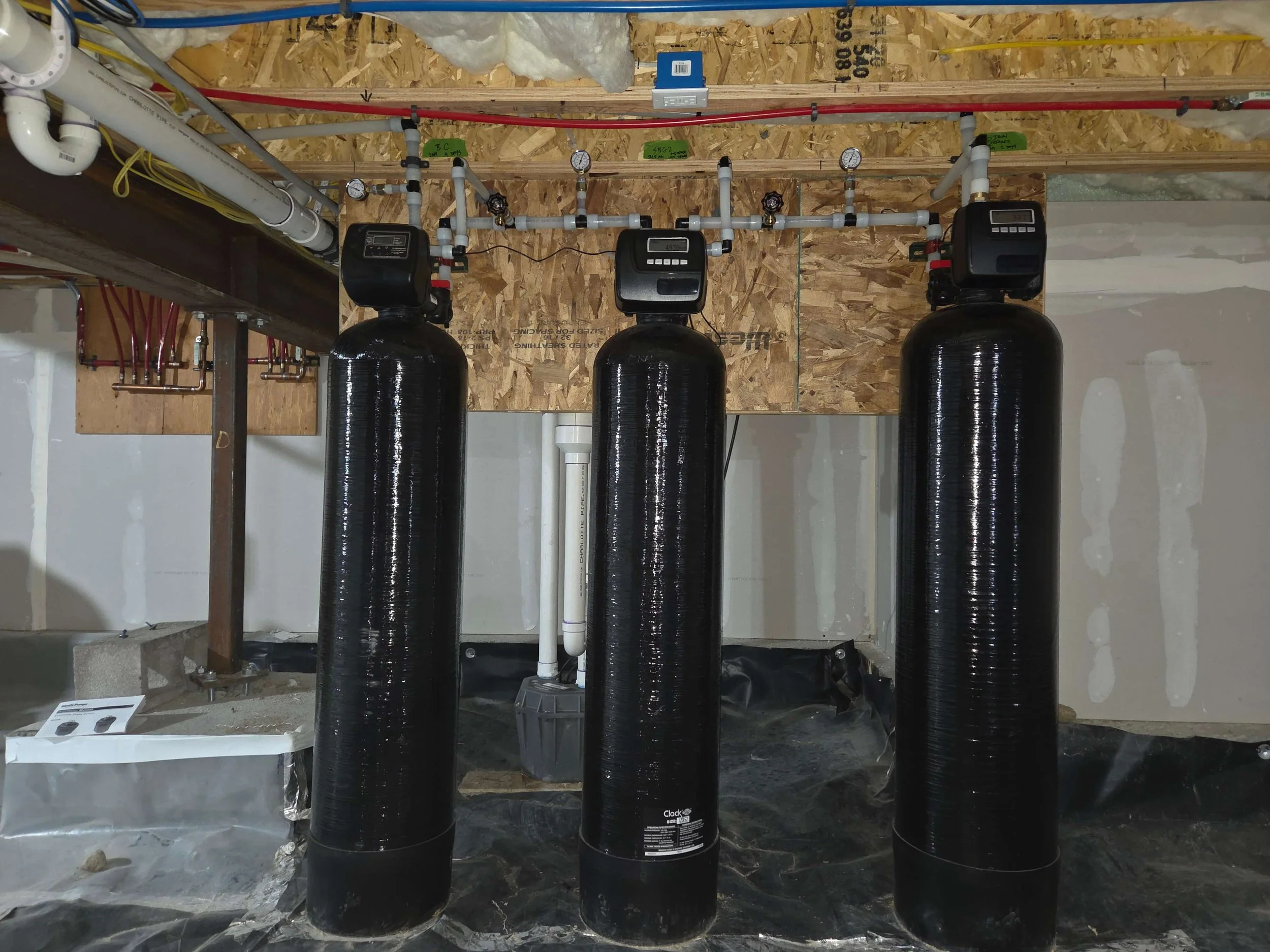 Three black water softener tanks with electronic control heads installed in a utility room, surrounded by plumbing, electrical wiring, and insulation on unfinished walls.