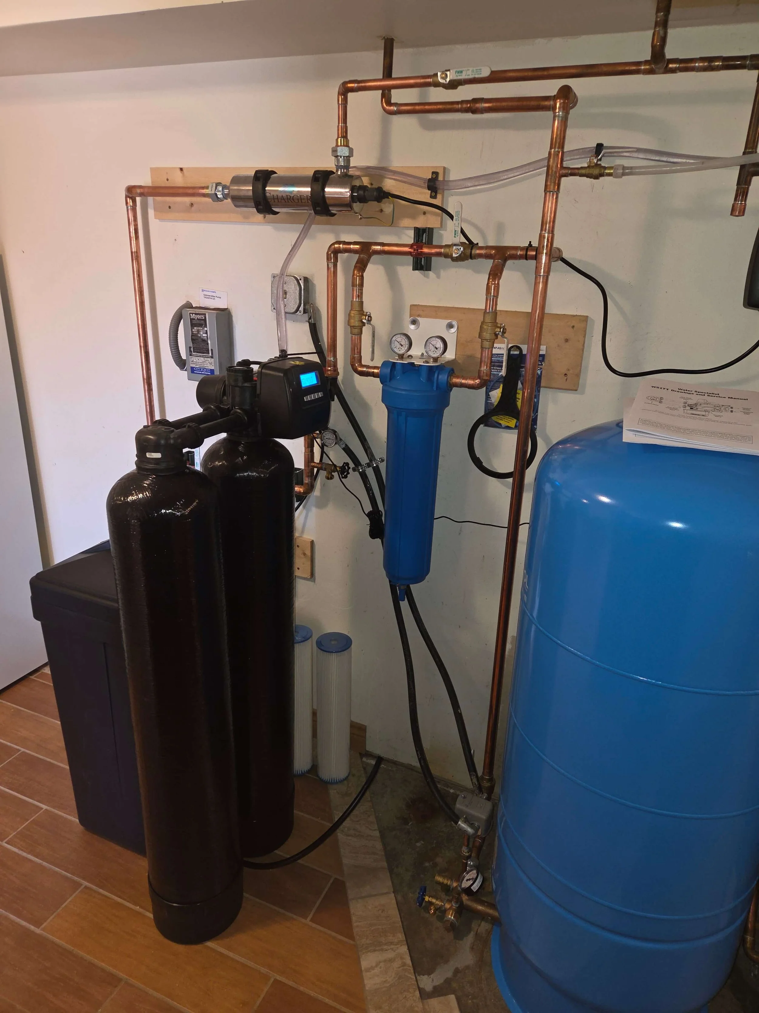 A water filtration or softening system with copper pipes, two black filtration tanks, a blue water softener tank, and gauges and controls mounted on a wall in a utility room.