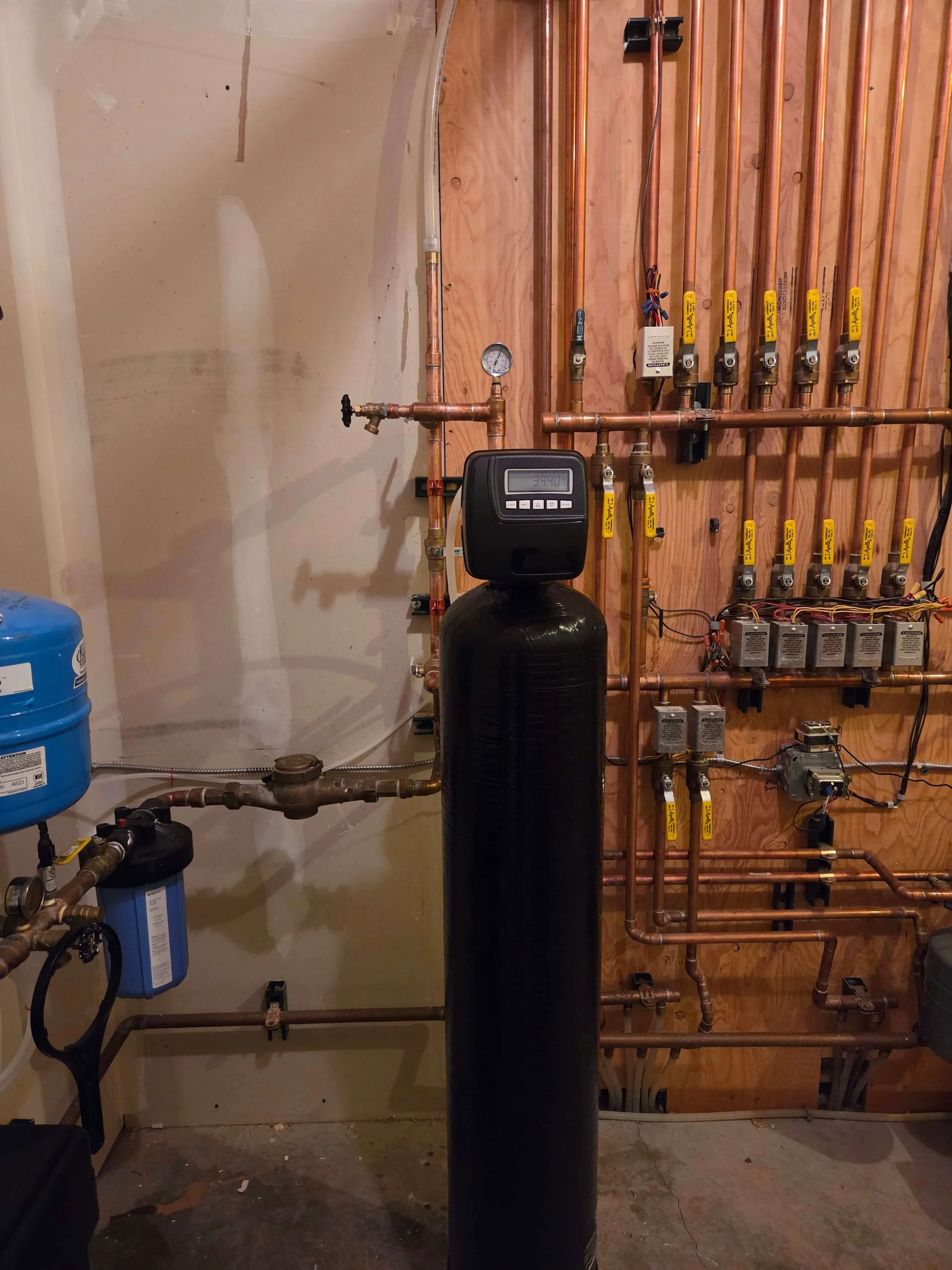 A plumbing or HVAC system setup with copper pipes, a black water filter tank, a digital control valve system, and various gauges and valves mounted on a wooden wall. The setup appears to be part of a water treatment or heating system.