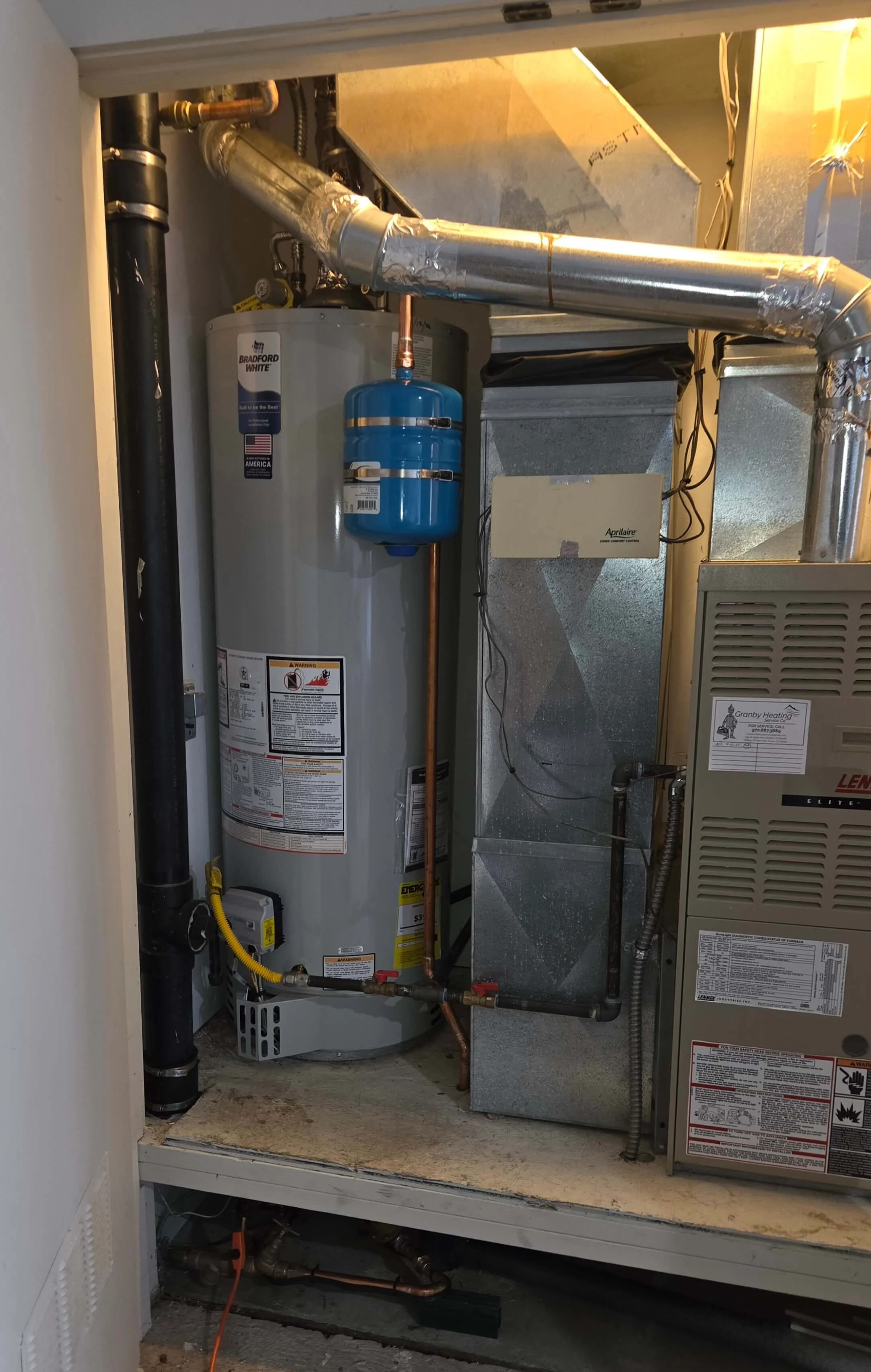 A utility closet containing a water heater with a blue expansion tank attached, insulated ductwork, a furnace, and various pipes and electrical wiring.