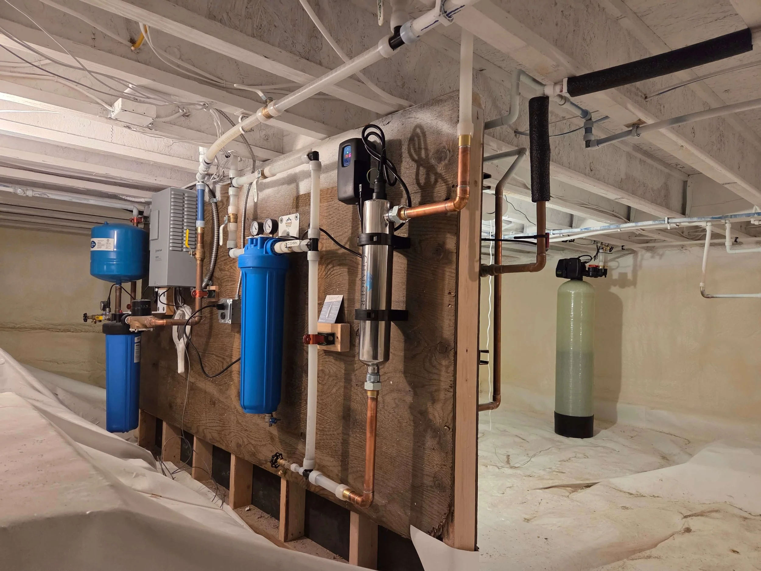 Plumbing and filtration system setup in a basement with pipes, water filters, gauges, and a pressure tank.