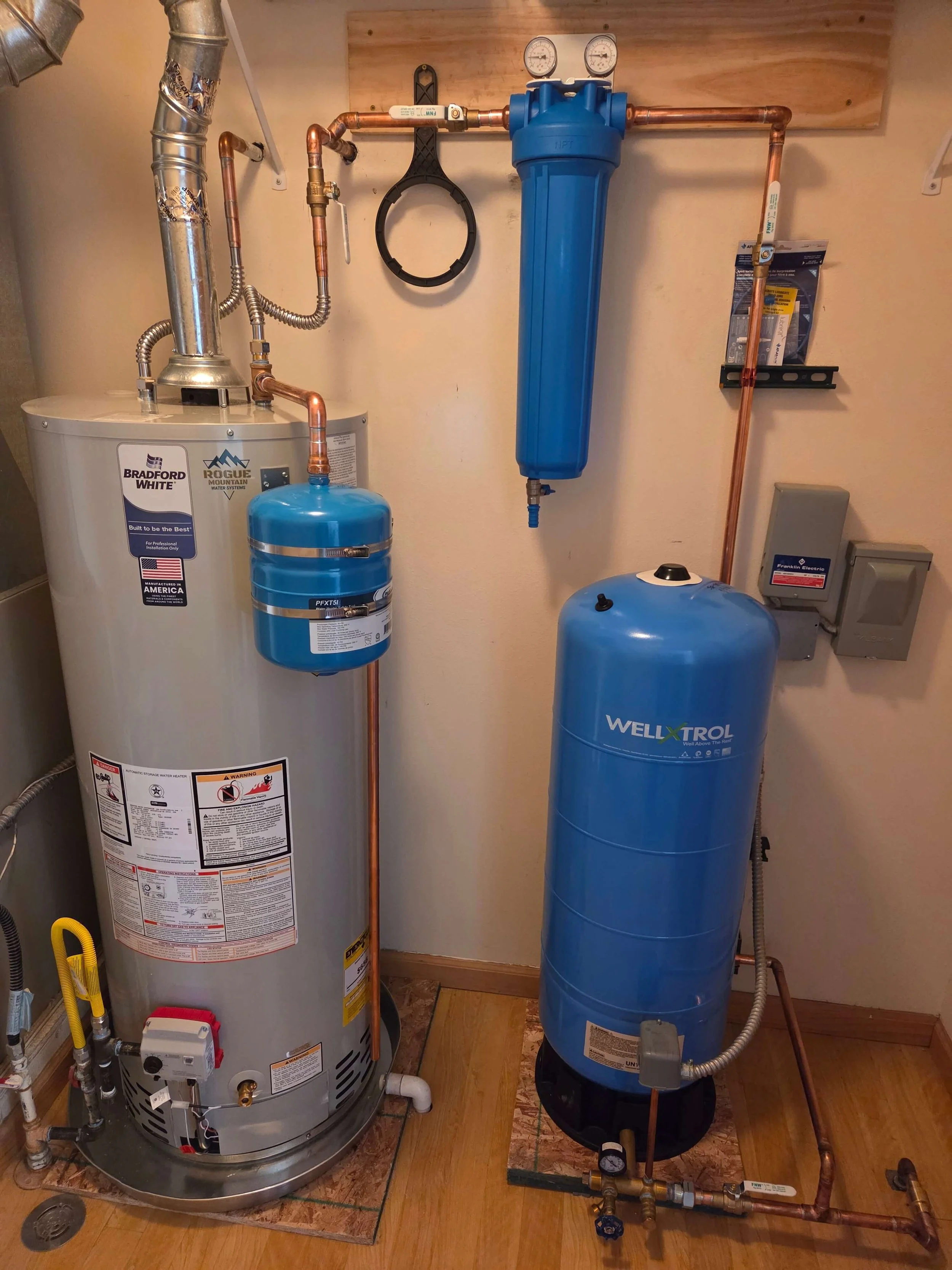 Water heater on the left with copper piping and a blue water filtration or softening unit on the right with additional piping, gauges, and control systems.