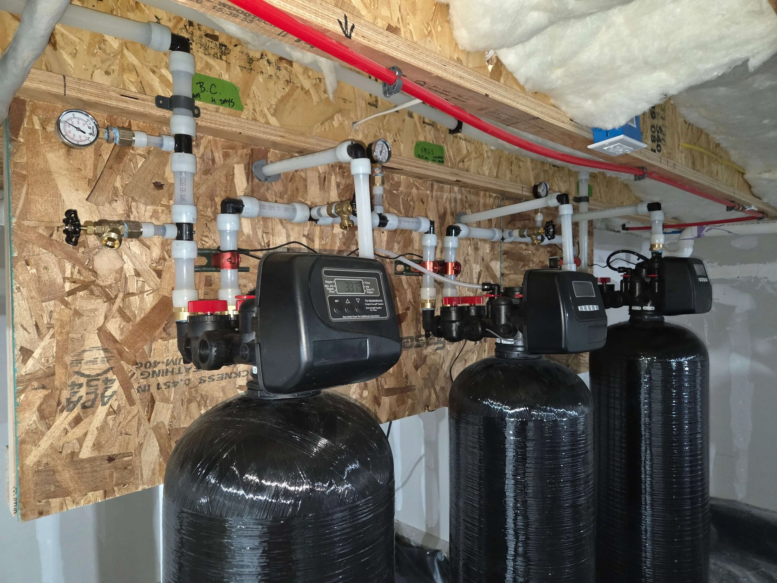 A water treatment system with multiple black cylindrical tanks, white PVC pipes, and two black control units installed on a wooden wall with insulation above.