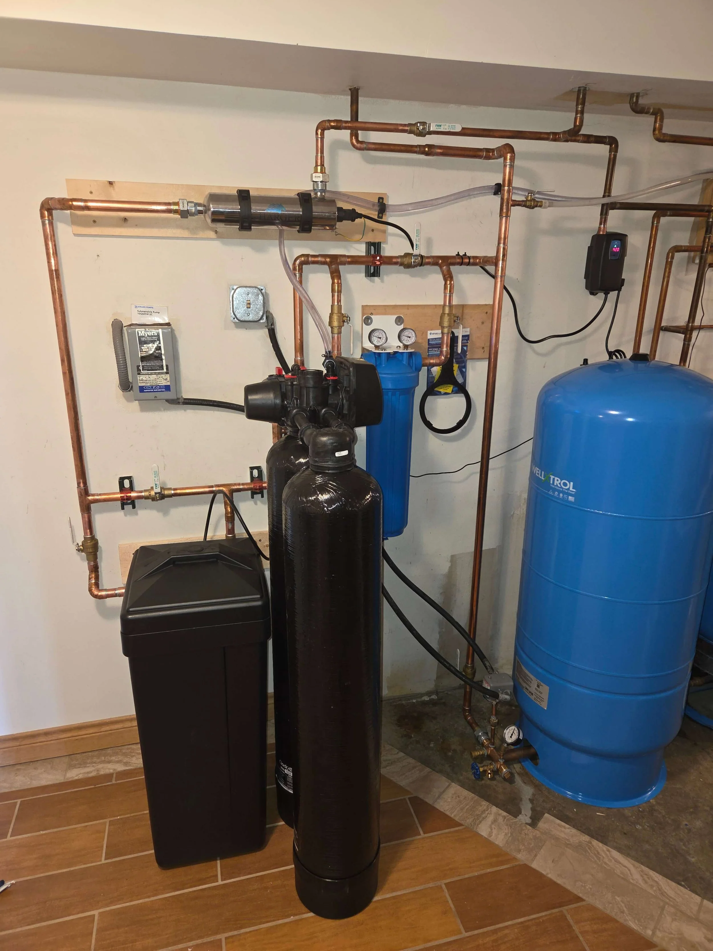 A water filtration system with copper pipes, a black water filter tank, a blue water softener tank, and gauges, installed in a basement.
