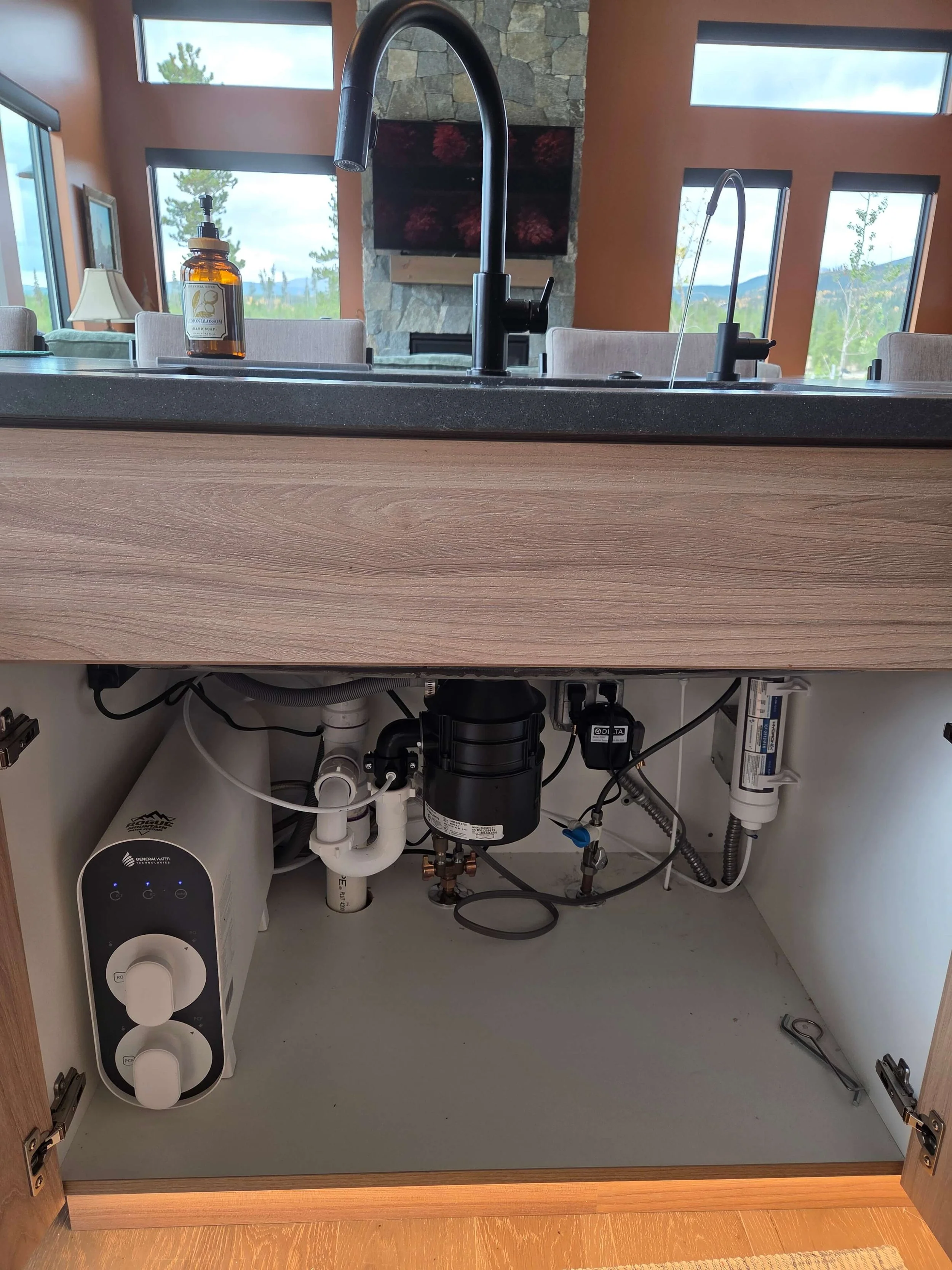 View of kitchen cabinet space with cabinet doors open, exposing plumbing and water filtration system underneath the sink, including a black water filter, white piping, and a water softener unit, with a gray countertop and wood flooring.