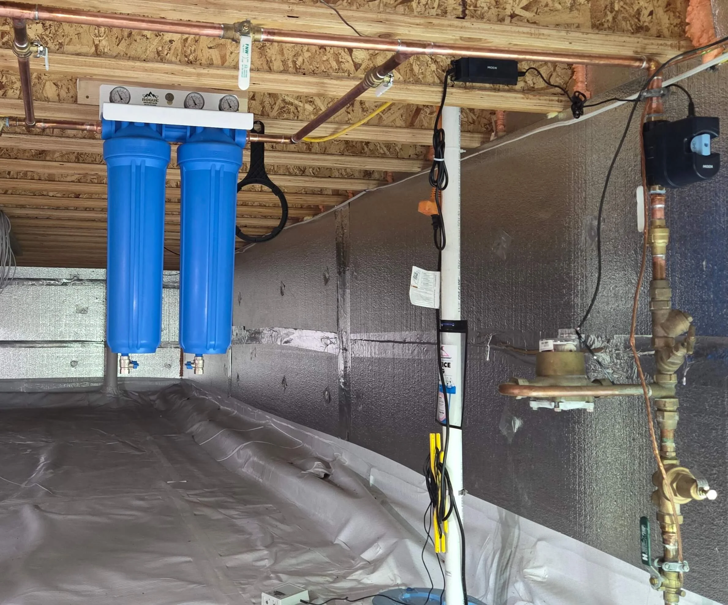 A plumbing setup with blue water filters connected to copper pipes, gauges, a water pump, and electrical wiring against a partially constructed wall with wooden framing.