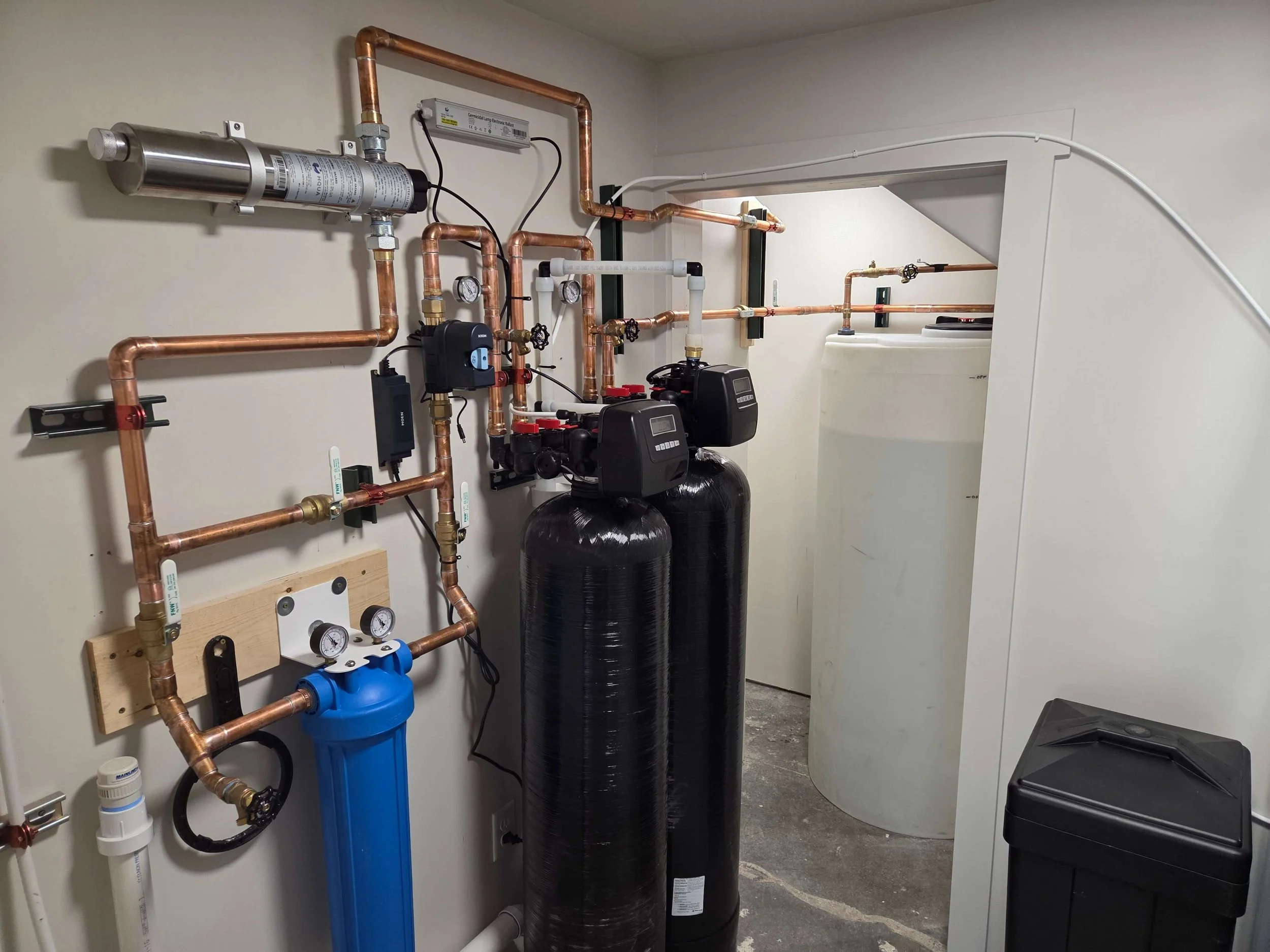 Interior view of a water filtration and softening system with copper pipes, two large black tanks, a blue water filter, gauges, and control units.