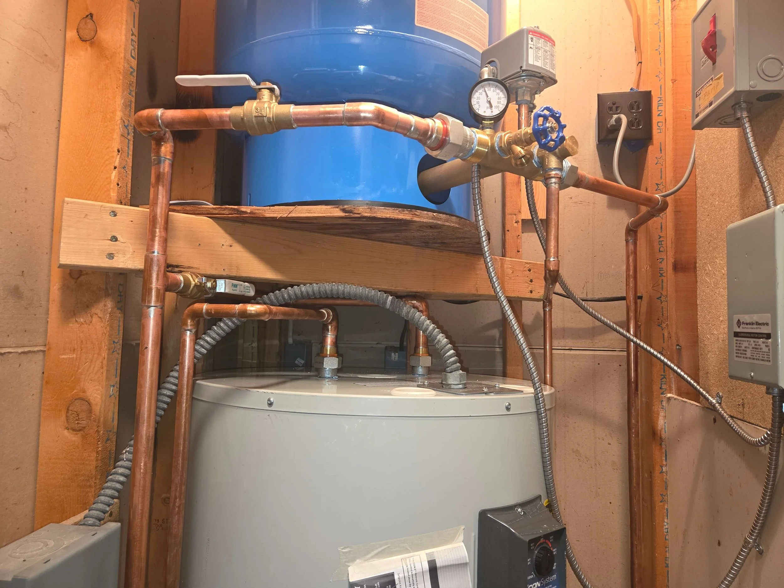 An electrical water heater with copper pipes, a pressure gauge, and a control module. There is a large blue tank in the background, mounted on a wooden frame with electrical wiring connected.