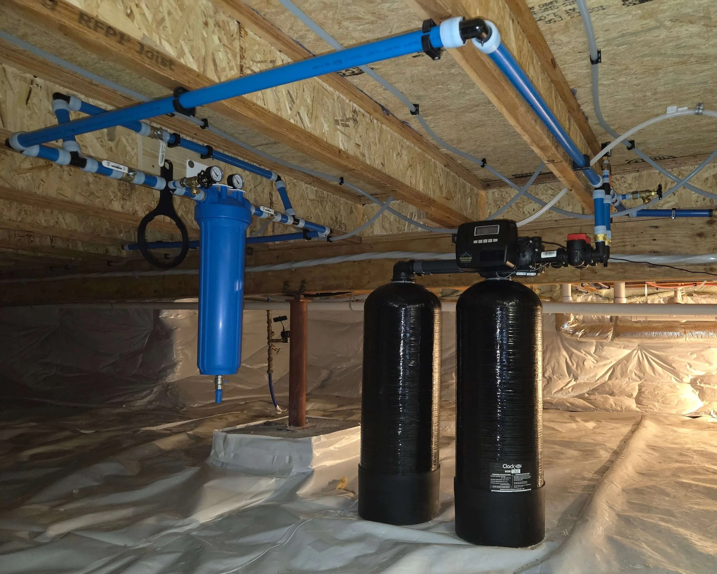 Underground plumbing pipes including blue water supply pipes and black water drain pipes, along with pressure gauges and a control valve, installed on a wooden ceiling with insulation.