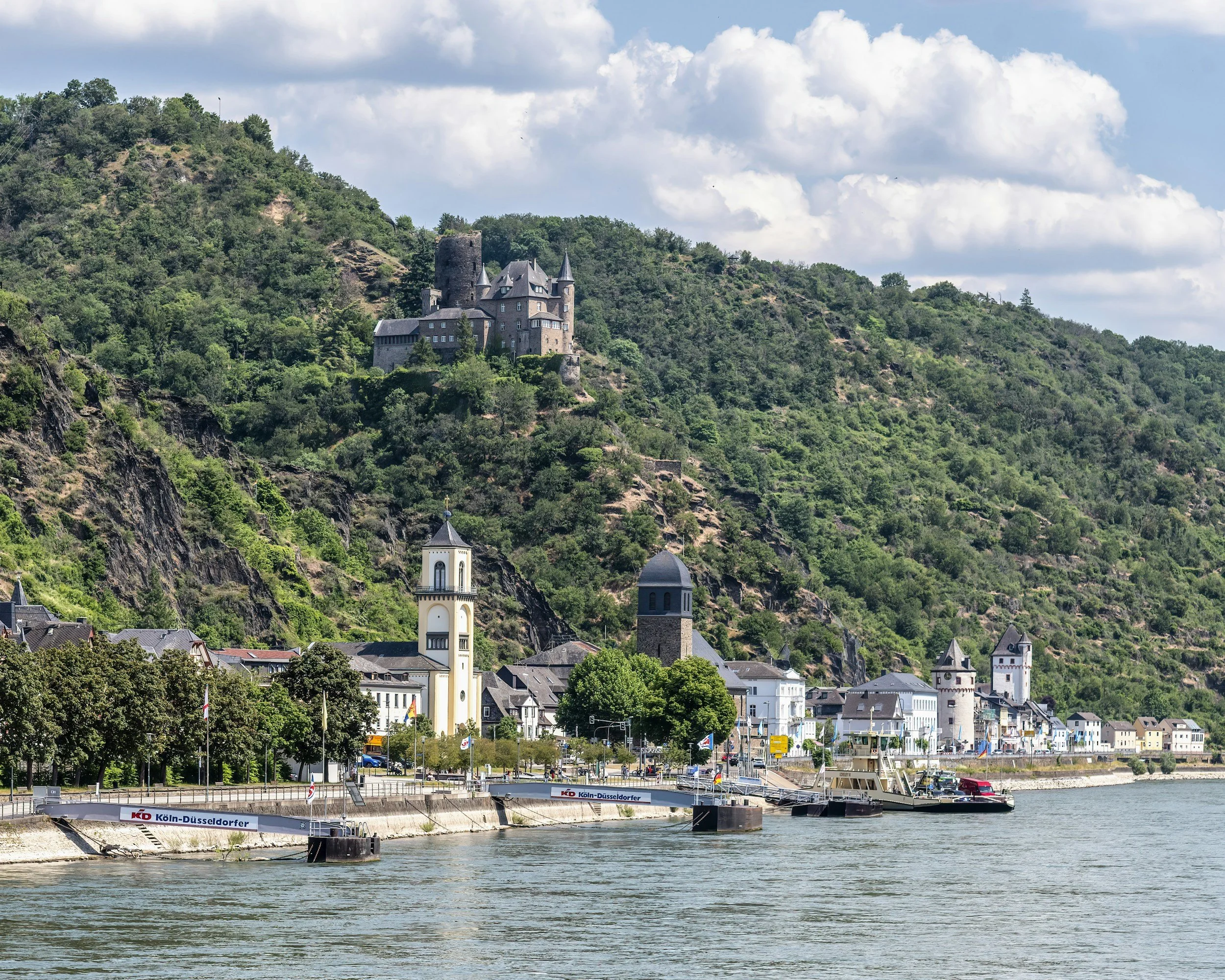 A scenic view of the Rhine River winding past a historic medieval castle on a lush hillside, representing the April 2026 guided wine cruise hosted by Kimber Travis.