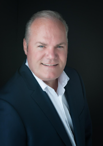 A professional headshot of a middle-aged man with gray hair, wearing a navy blue blazer and a white shirt, smiling against a dark background.