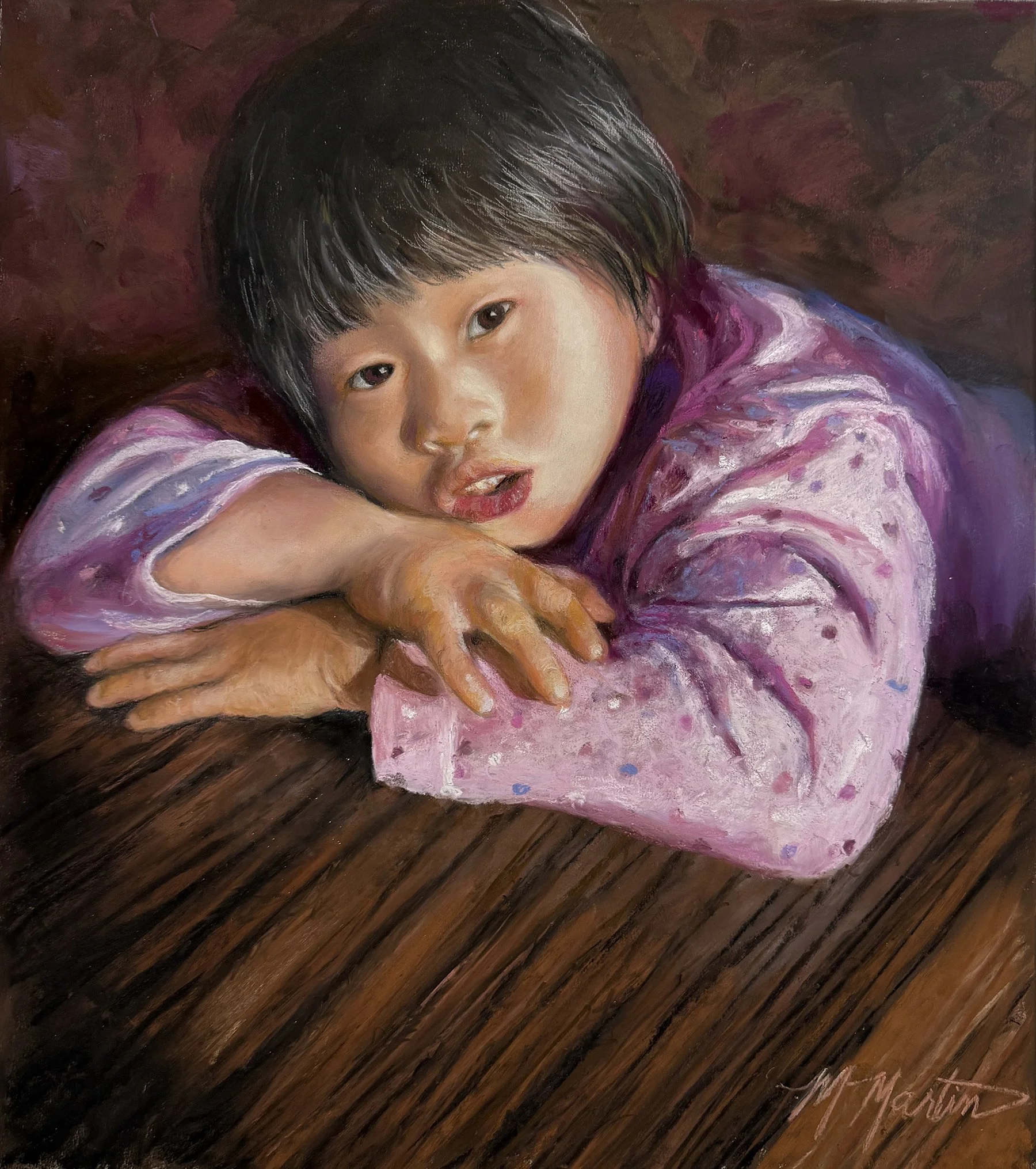A young girl with short black hair resting her head and arms on a wooden surface, wearing a pink and purple polka-dotted long-sleeve shirt, looking directly at the camera.