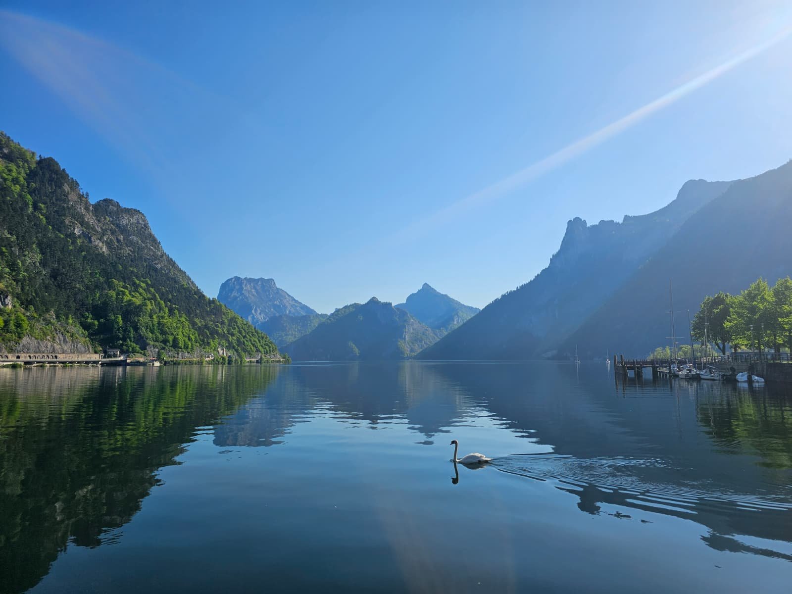 Ein See mit Bergen im Hintergrund, ein Schwäne schwimmt im Wasser, und es gibt Boote am Ufer auf der rechten Seite, bei hellem Sonnenlicht.