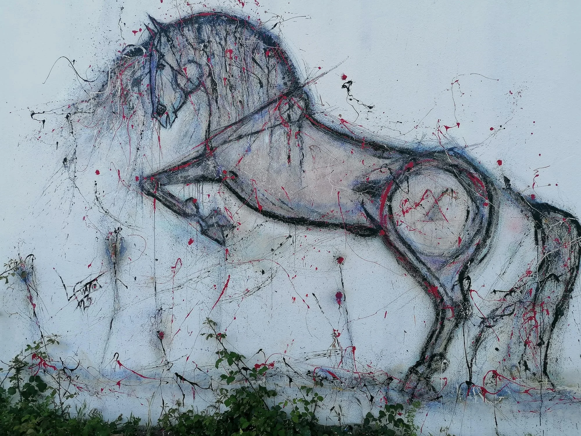 Grafite de um cavalo em uma parede branca, feito com técnica de pintura splatter com tinta preta, branca, vermelha e roxa, com detalhes de folhagens na parte inferior.