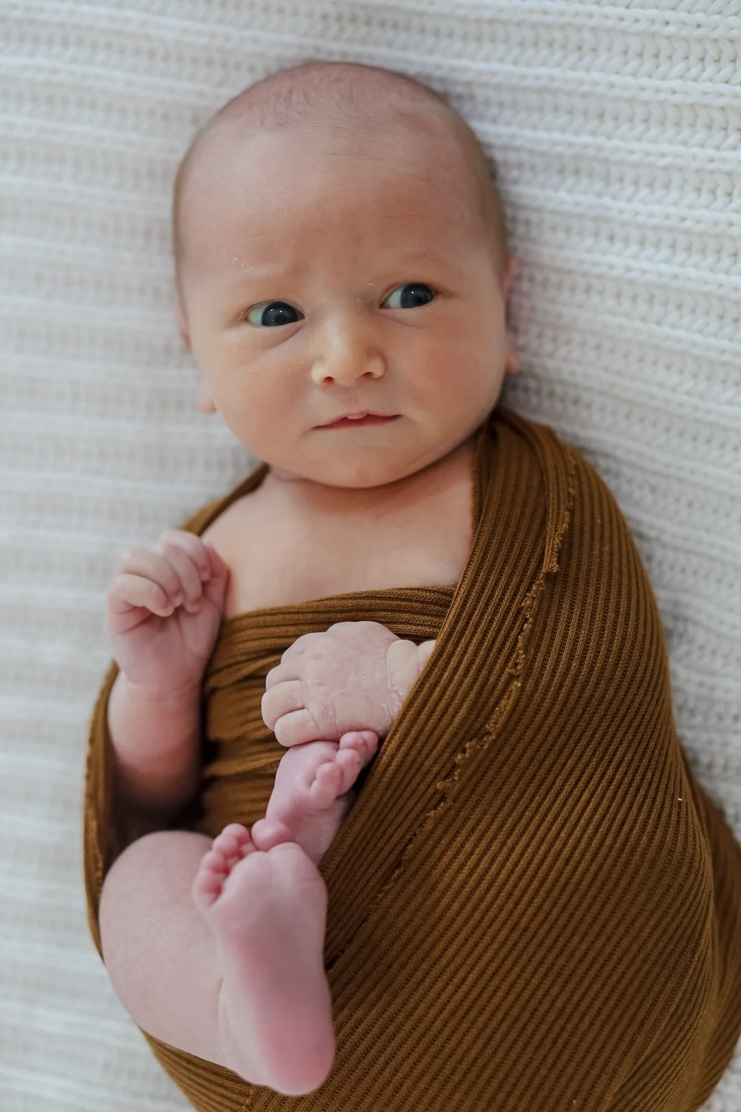 Newborn baby wrapped in brown blanket lying on a white textured background.