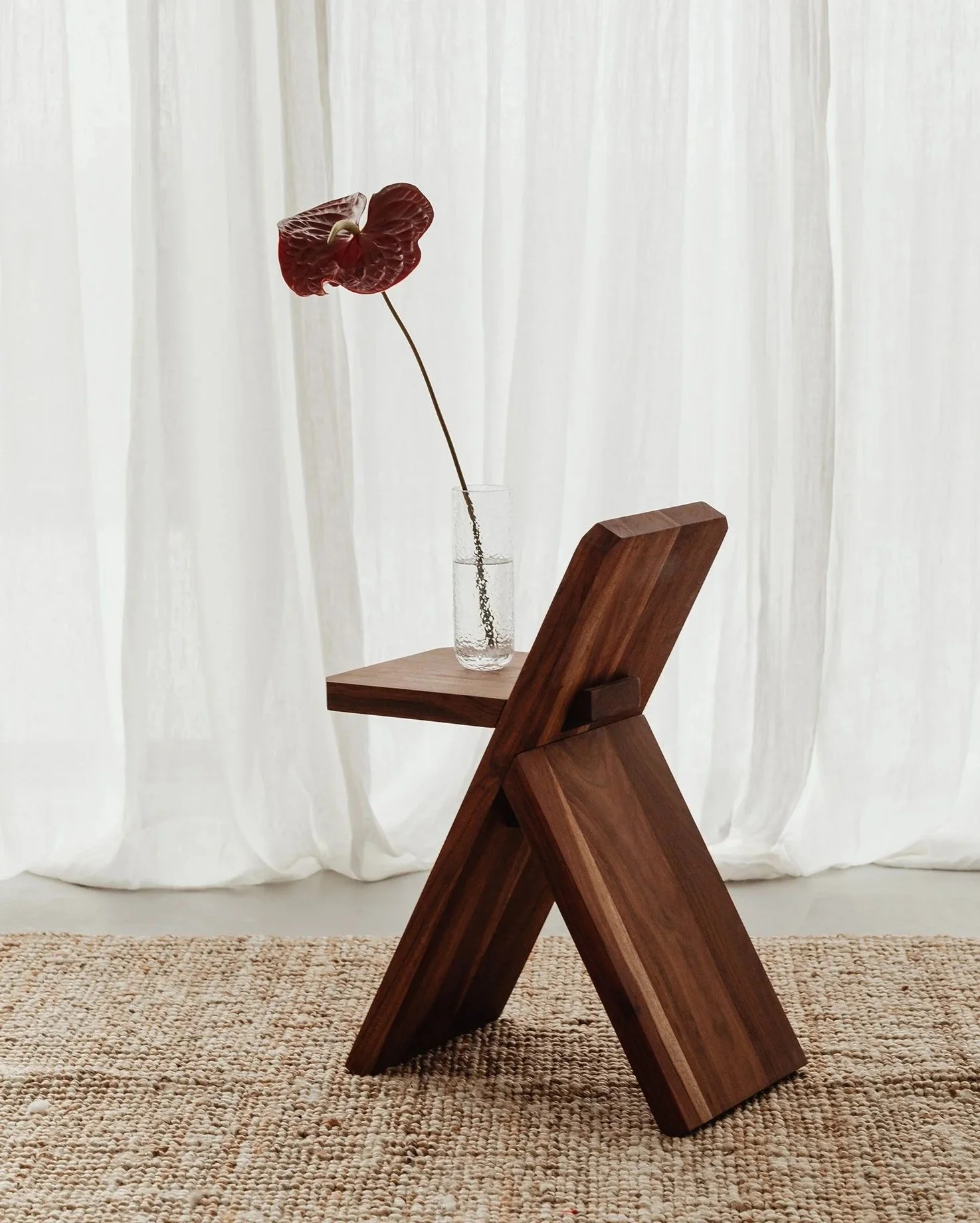 A wooden chair with an angular design, a glass vase with a single dark red anthurium flower, set against a backdrop of white curtains and a woven beige rug.