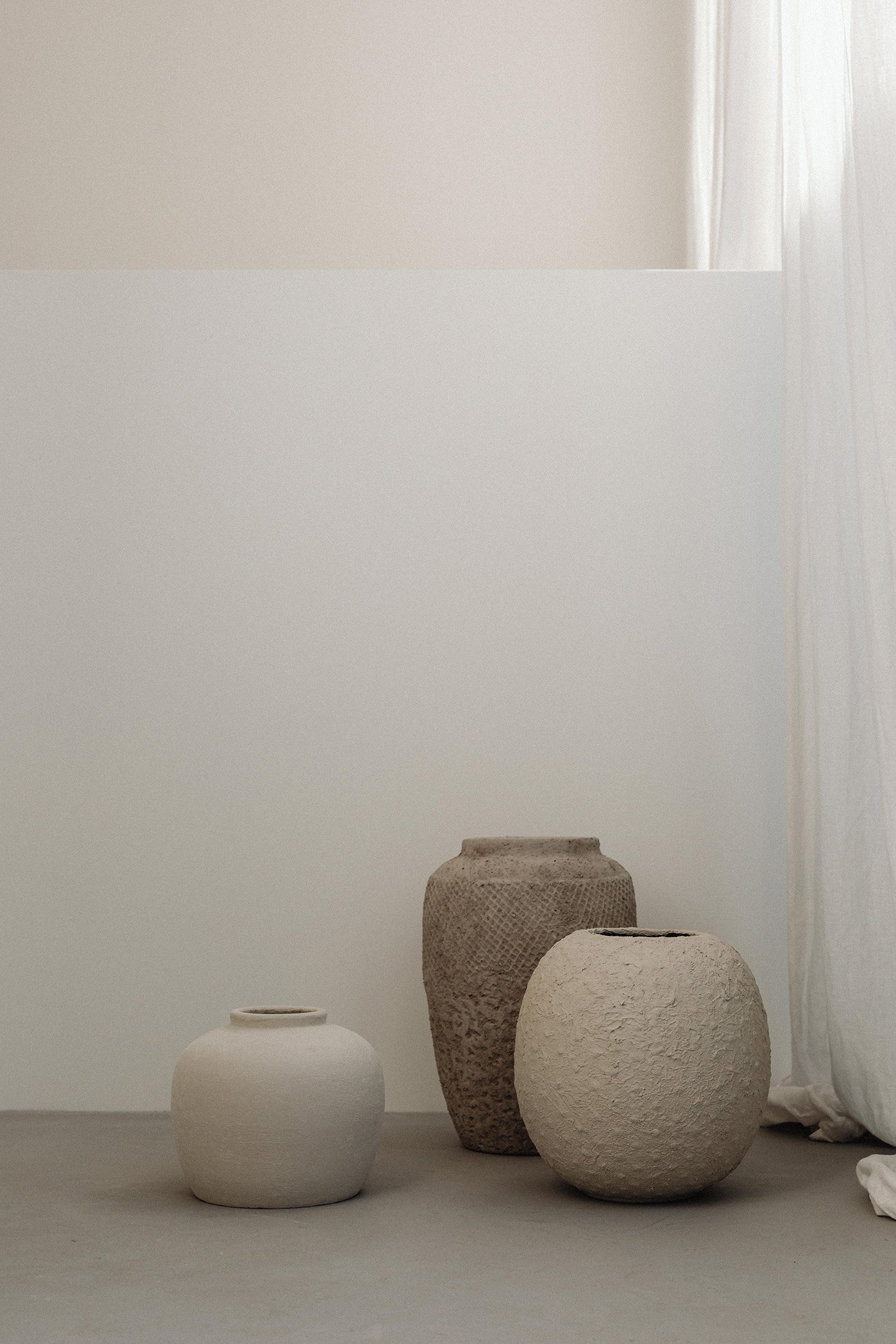 Three minimalistic pottery vases of different shapes and colors placed on a plain surface against a white wall, with part of a white curtain on the right side.