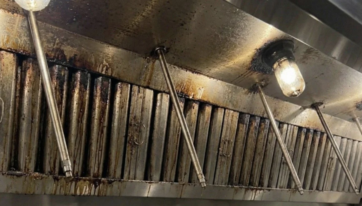 A greasy commercial kitchen exhaust hood with four sprinkler heads and a light bulb.