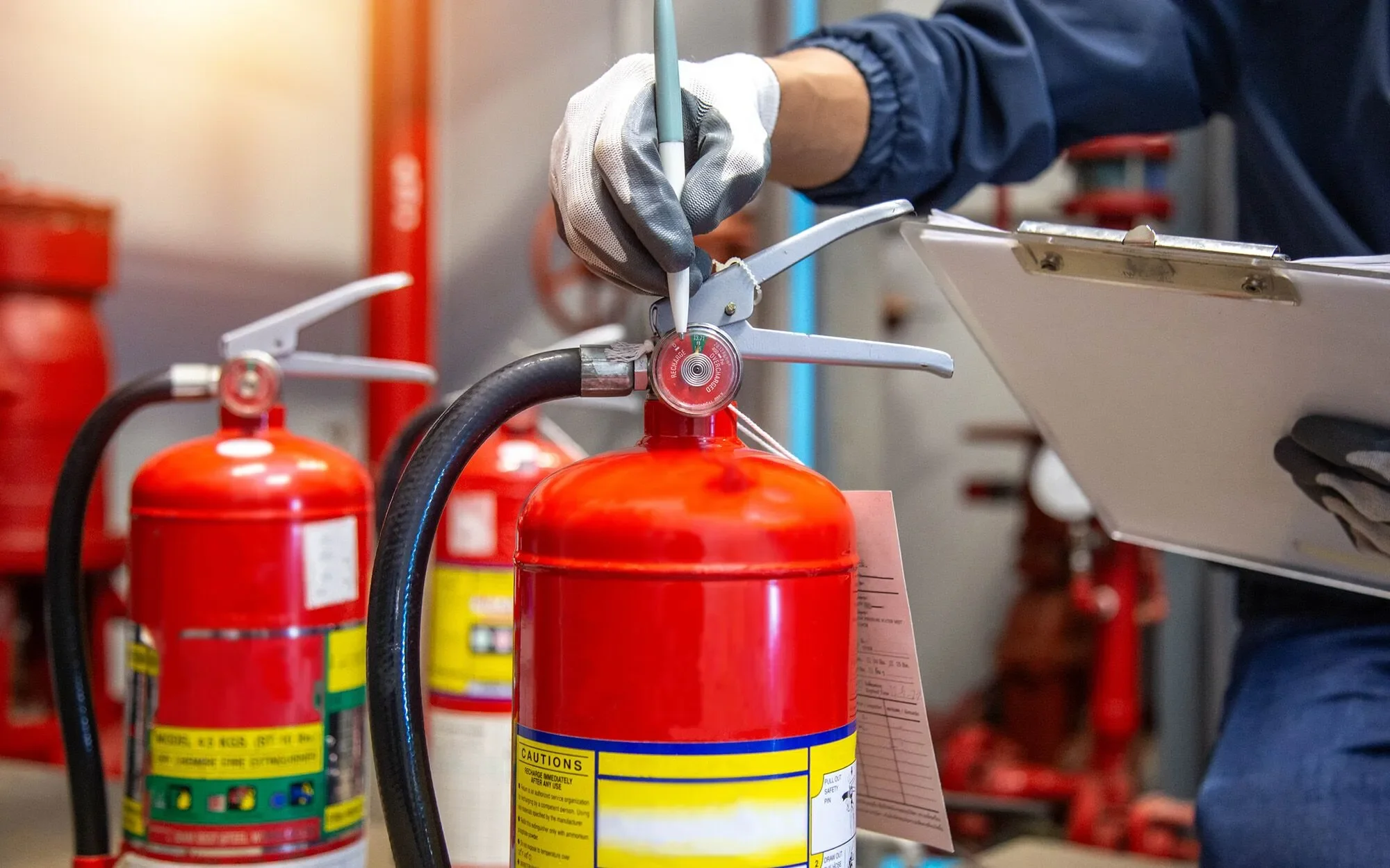 A person wearing gloves using a pen to write on a tag attached to a red fire extinguisher, with several other fire extinguishers in the background.