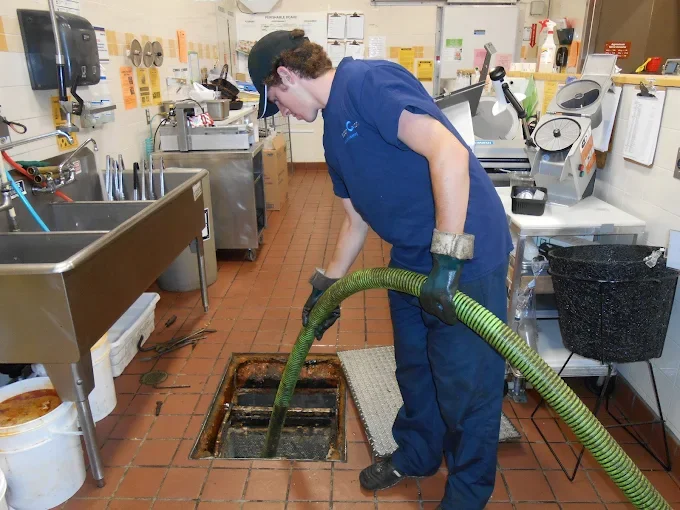 A worker in a blue uniform and gloves cleaning a greasy kitchen drain with a large green hose in a commercial kitchen or restaurant.