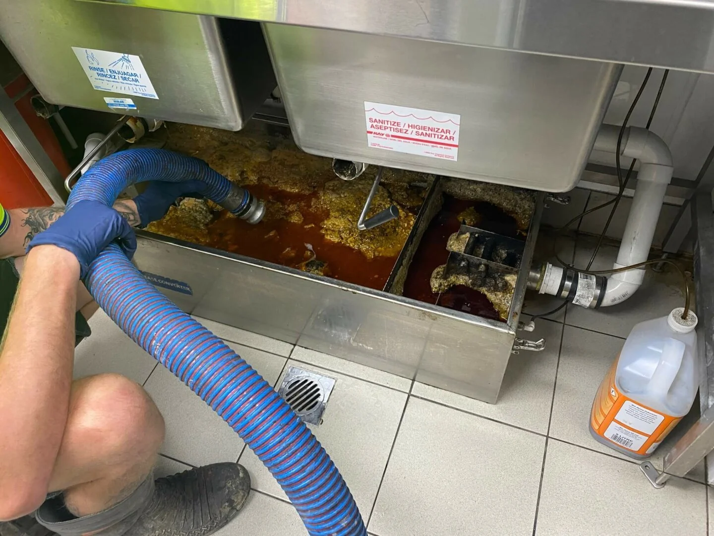 A person wearing blue gloves and black boots is cleaning a grease trap with a large blue hose. The grease trap is filled with dirty, greasy, and brownish liquid, with some areas of accumulated solid waste. There are warning signs on the grease trap, and a bottle of cleaning solution on the floor nearby.