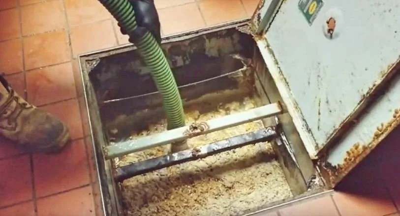 Open hatch in the floor revealing a sump pump with a green hose, plumbing inside a kitchen or utility room with tiled floor and a person standing nearby.