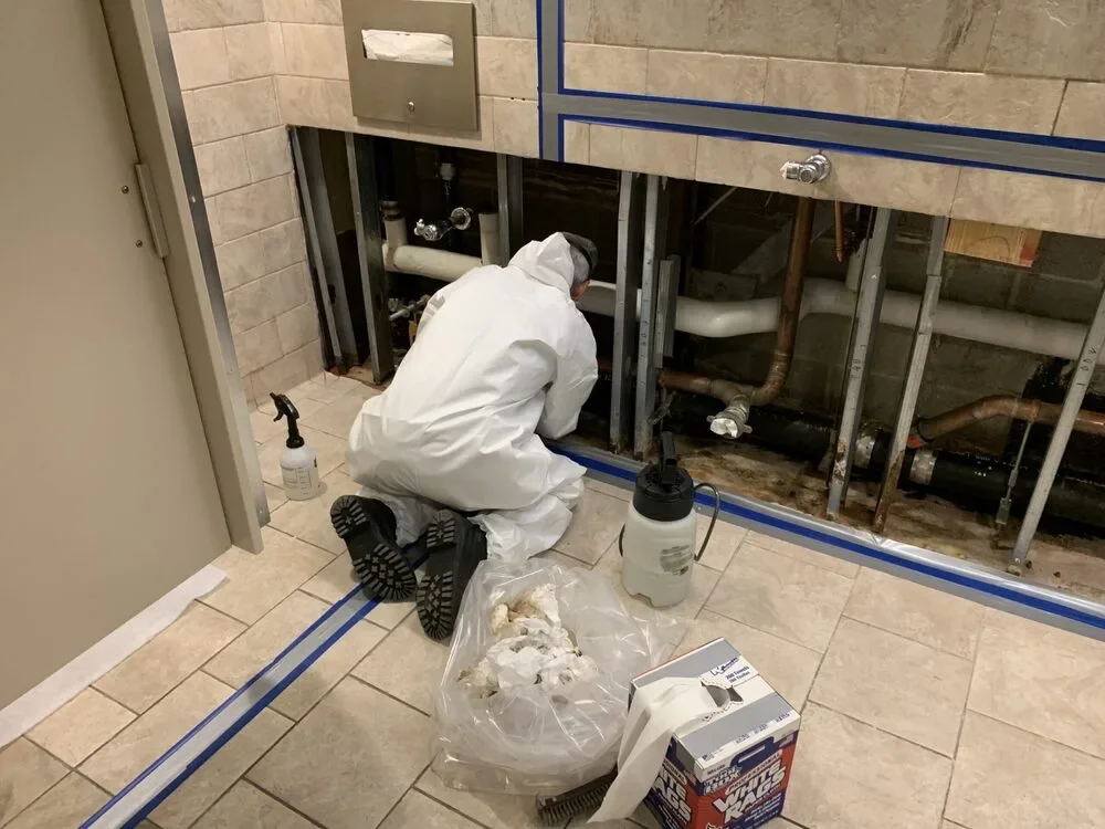 A worker in protective gear repairing plumbing underneath a bathroom sink. Various tools and supplies are on the floor.