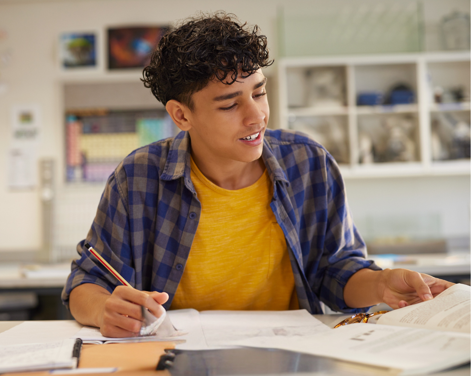 A young man wearing a yellow t-shirt and blue plaid shirt at a desk in a classroom, doing an ACT/SAT practice test and holding a pencil.
