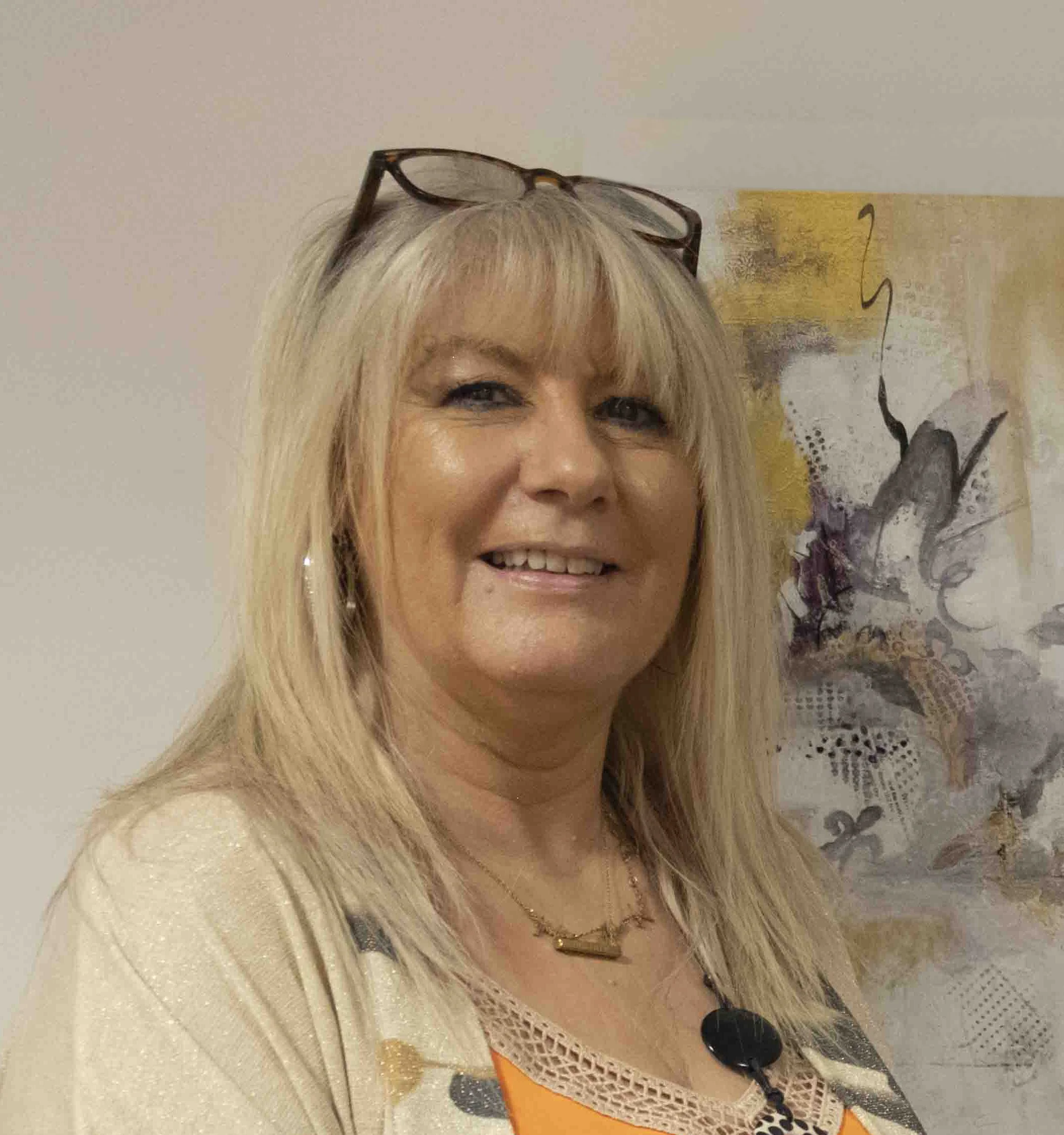 A smiling woman with blonde hair styled in a spiky fashion, wearing a colorful dress with abstract patterns, large leopard print jewelry, and earrings, standing in front of a light-colored wall and a framed artwork.