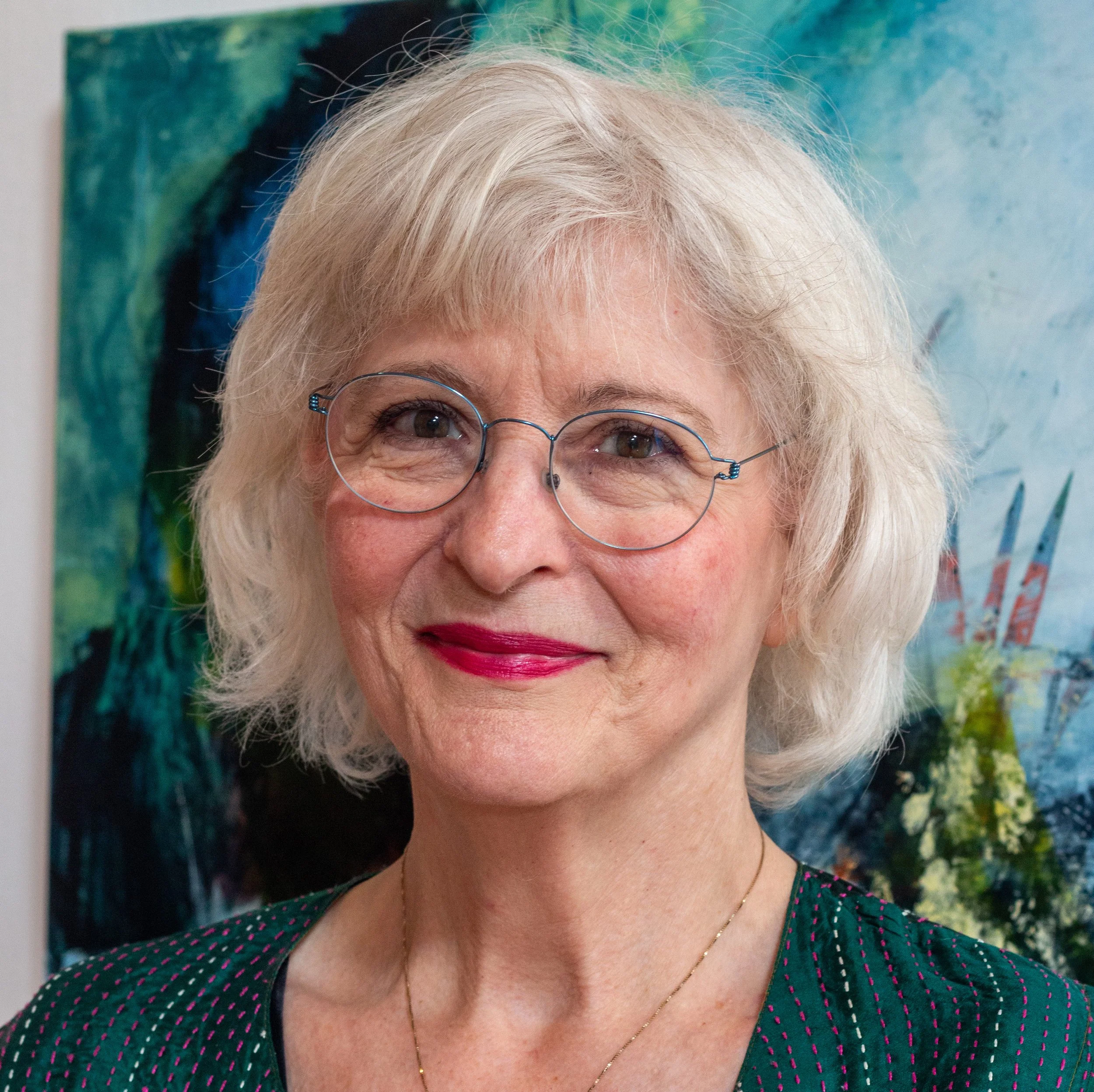 Smiling elderly woman with white hair, wearing a turquoise necklace and a light green top, standing outdoors by the water with rocky shoreline and mountains in the background.