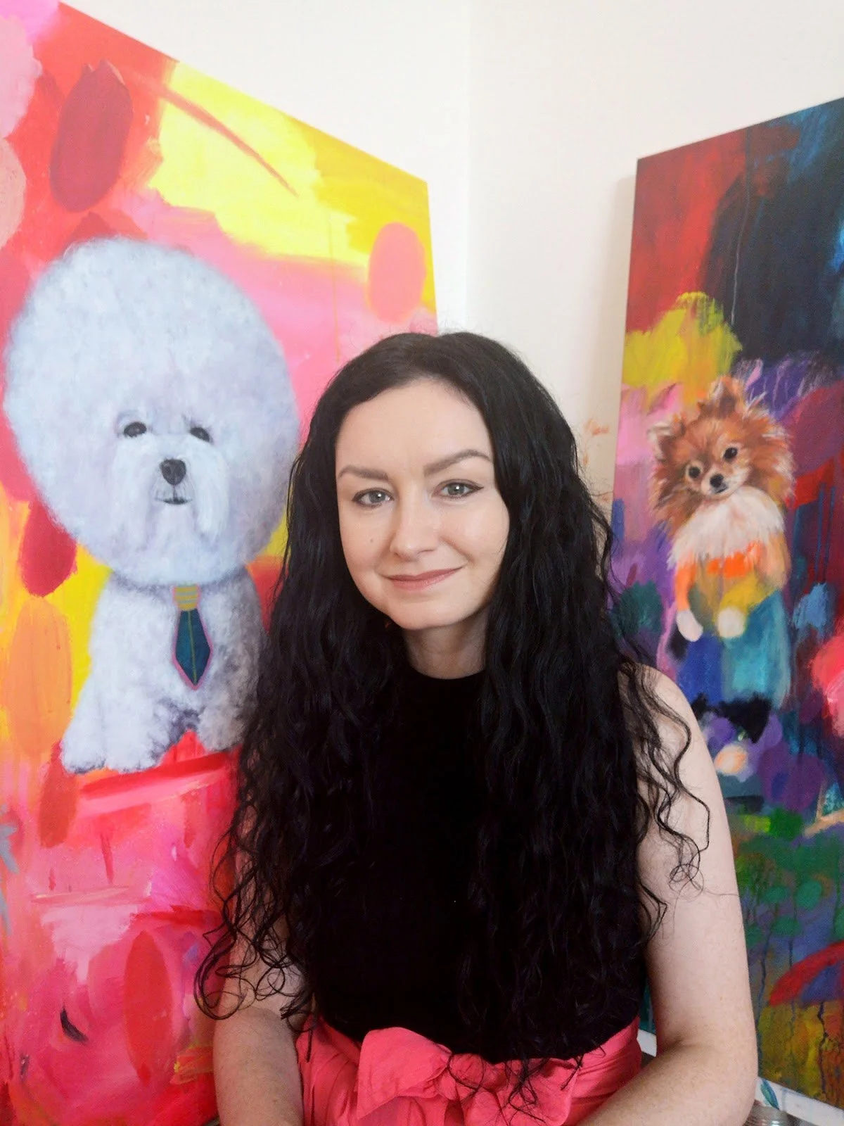 A woman with long, curly black hair and fair skin standing in front of colorful paintings of dogs.