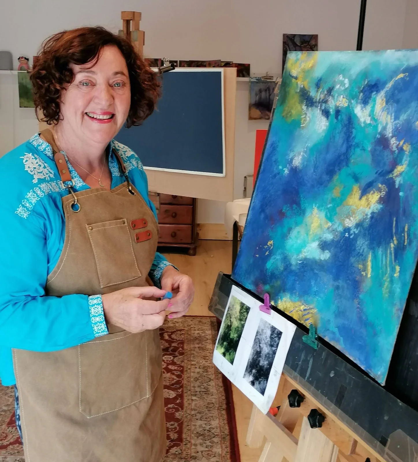 A woman with curly brown hair smiling at the camera while standing in front of an easel with a colorful abstract painting. She is wearing a blue shirt and a tan apron.