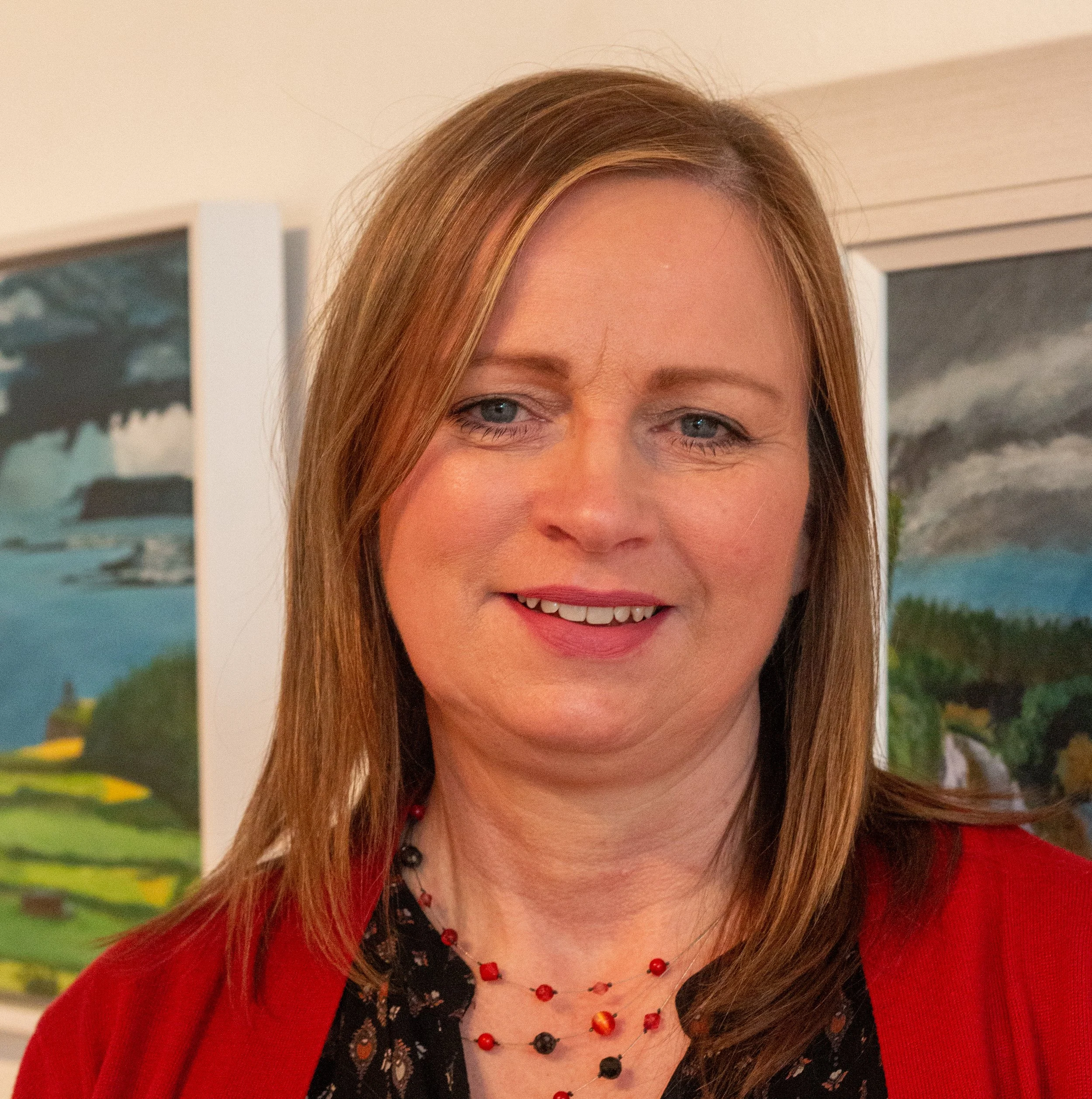 A woman with light brown hair smiling in front of a landscape painting of lavender fields on a gallery wall.