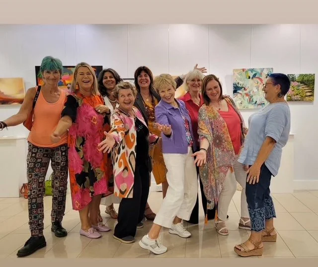 Group of women at an art gallery, smiling and posing in front of paintings.
