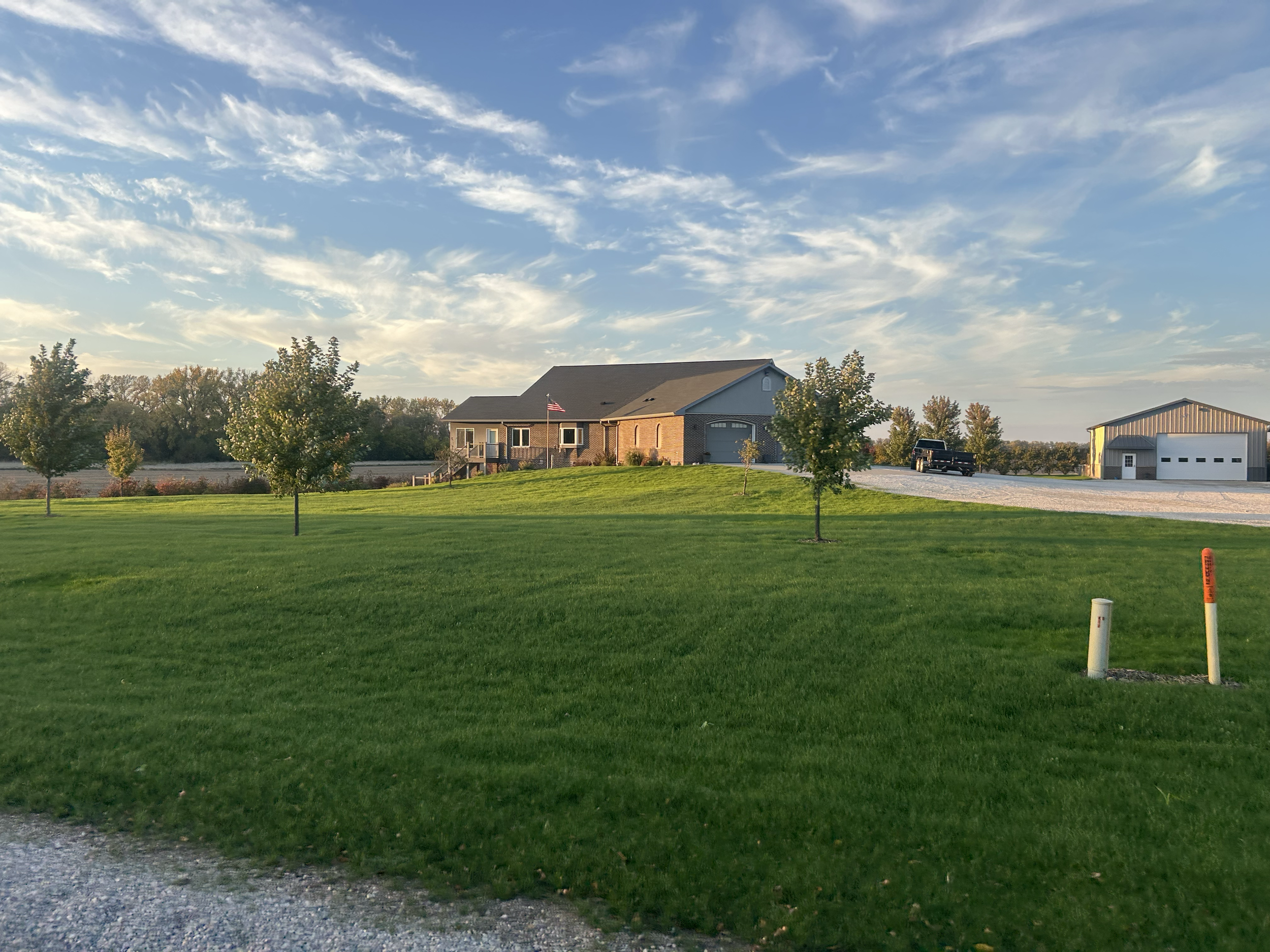 A house with a brick and siding exterior, a porch with steps, an American flag, a black truck parked on a gravel driveway, surrounded by a well-maintained lawn and small trees, with a barn in the background and a partly cloudy sky.