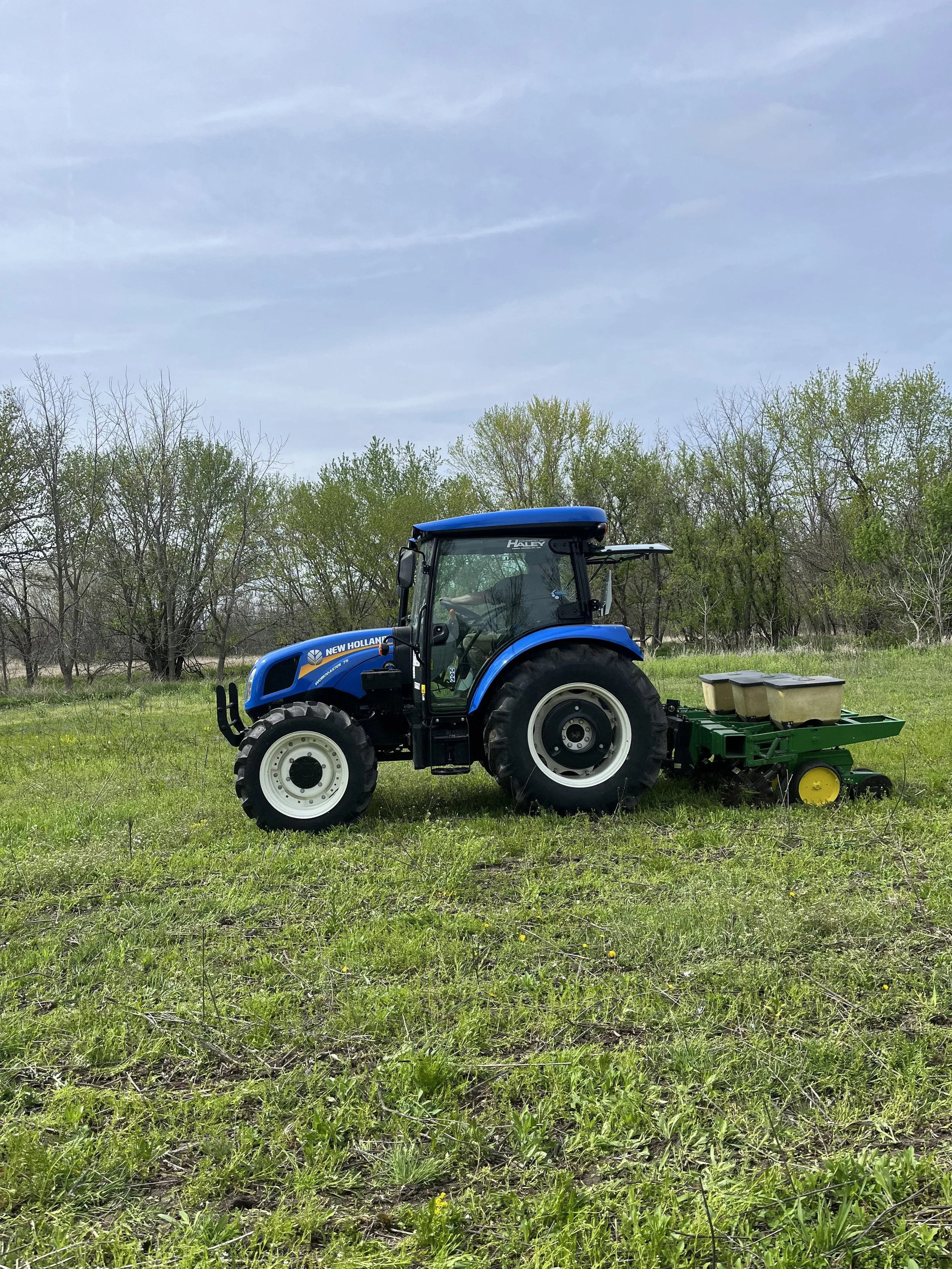 Blue New Holland tractor with a green planter attached, working on a grassy field during daytime with trees and a cloudy sky in the background.