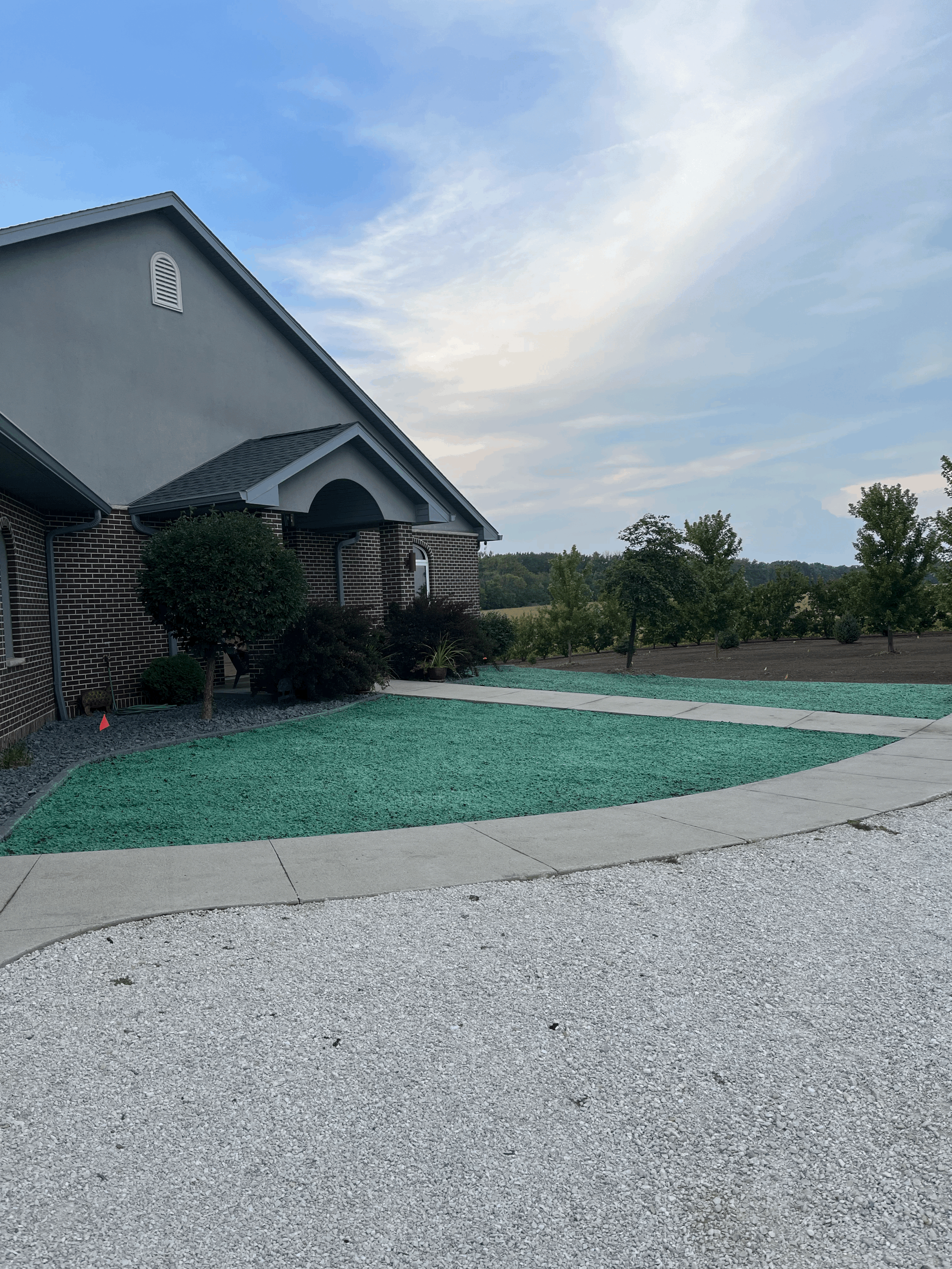 View of a residential house with a landscaped yard, sidewalk, and gravel driveway, under a partly cloudy sky.