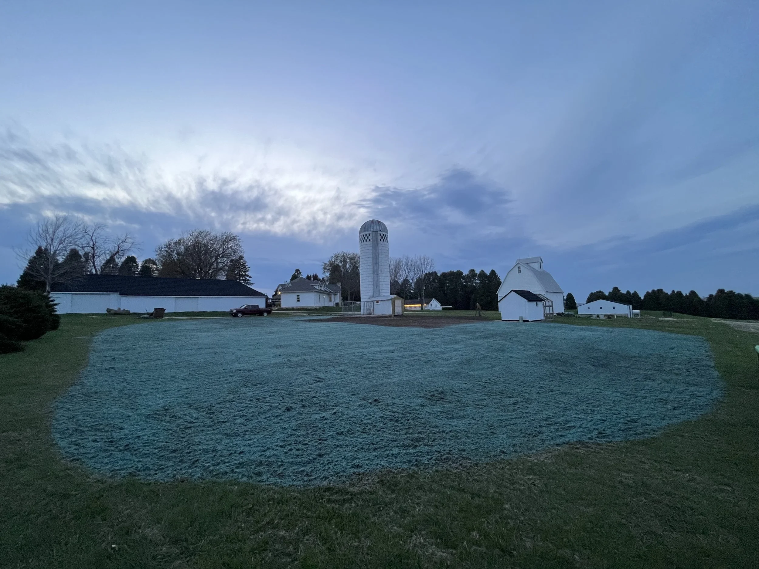 A farm scene at dusk with a white silo, several white barns, a black roofed building, a parked car, and trees in the background under a cloudy sky.