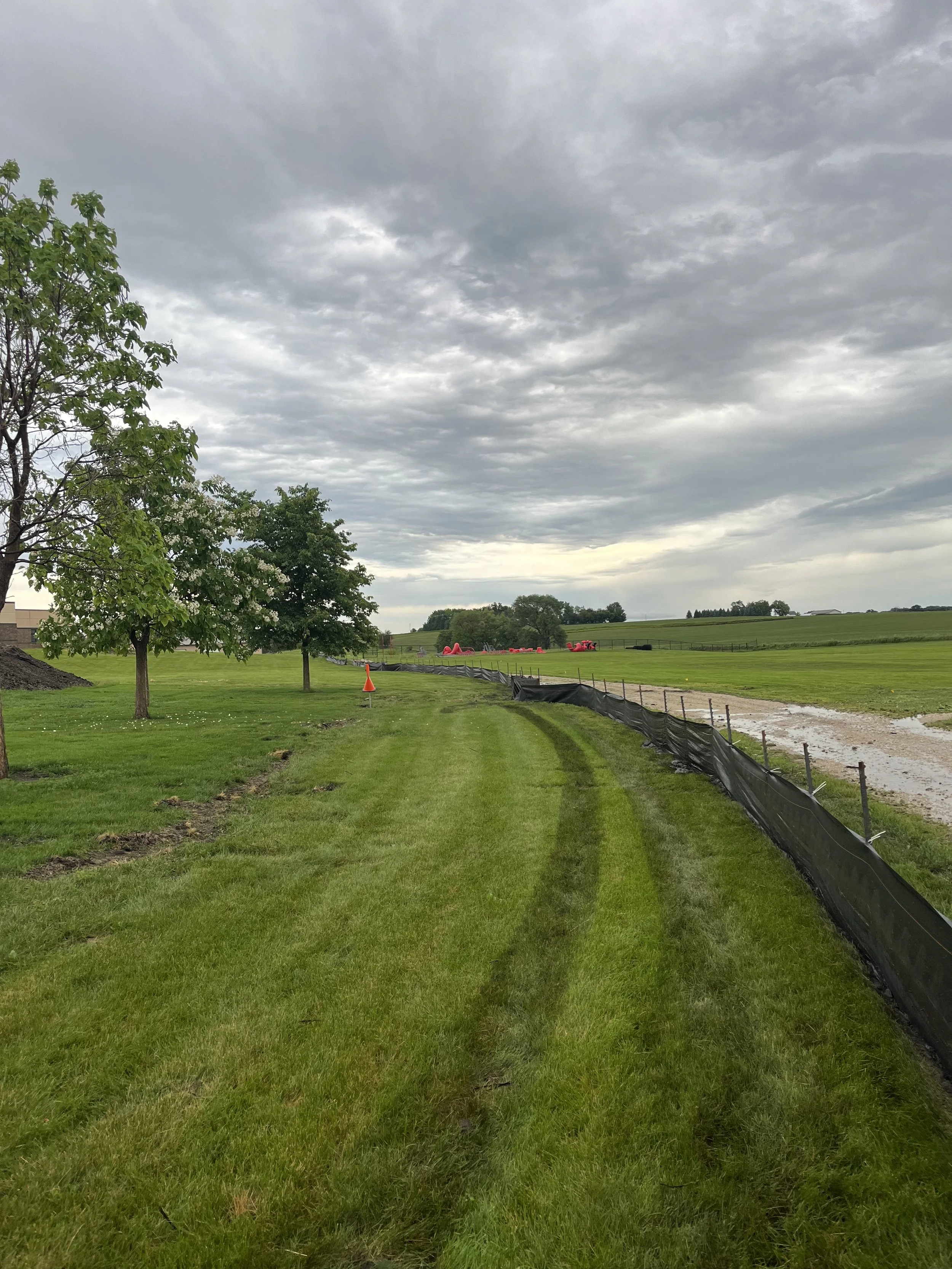 Green grassy field with tire tracks, trees, construction cones, and a dirt pathway under a cloudy sky.