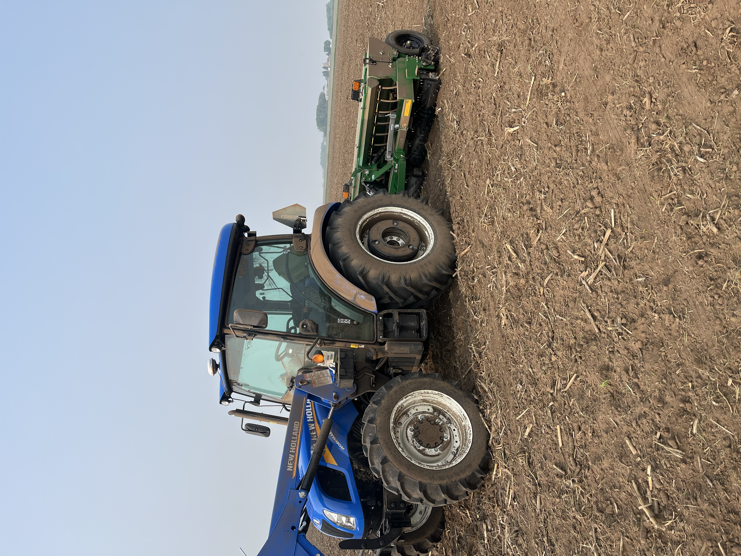 A blue New Holland tractor with large tires is parked on a freshly plowed field, with a green agricultural implement attached behind it.