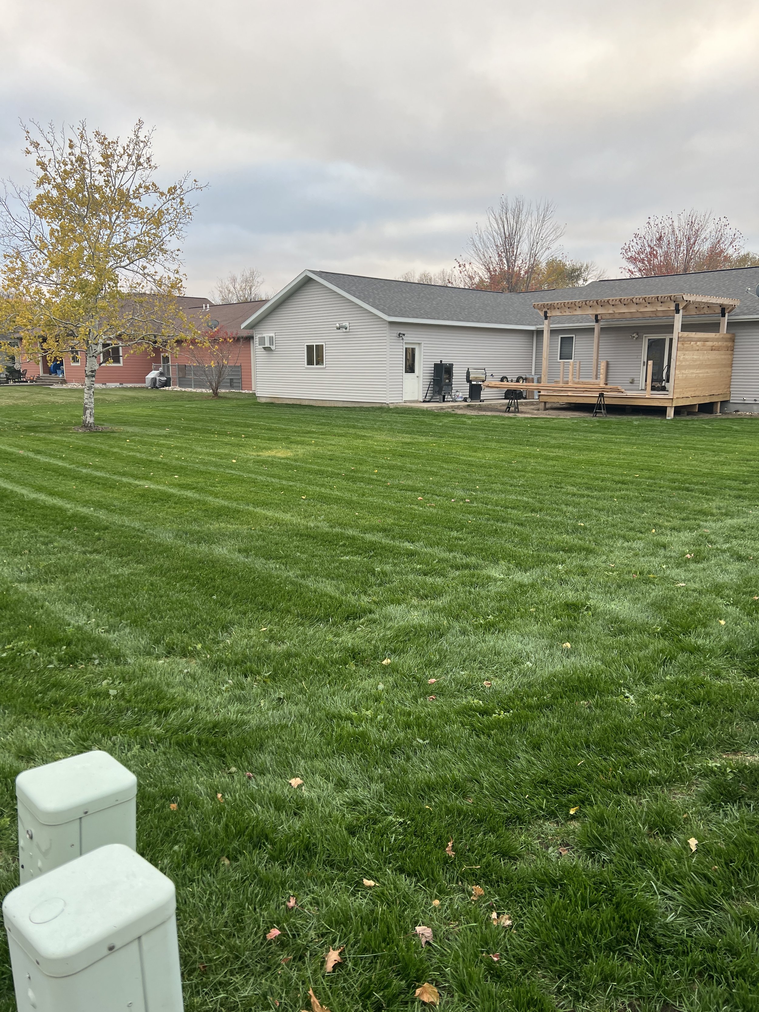 A backyard with neatly mowed green grass, a tree with yellowing leaves, a small house with gray siding, a wooden deck under construction, and two utility boxes in the foreground.