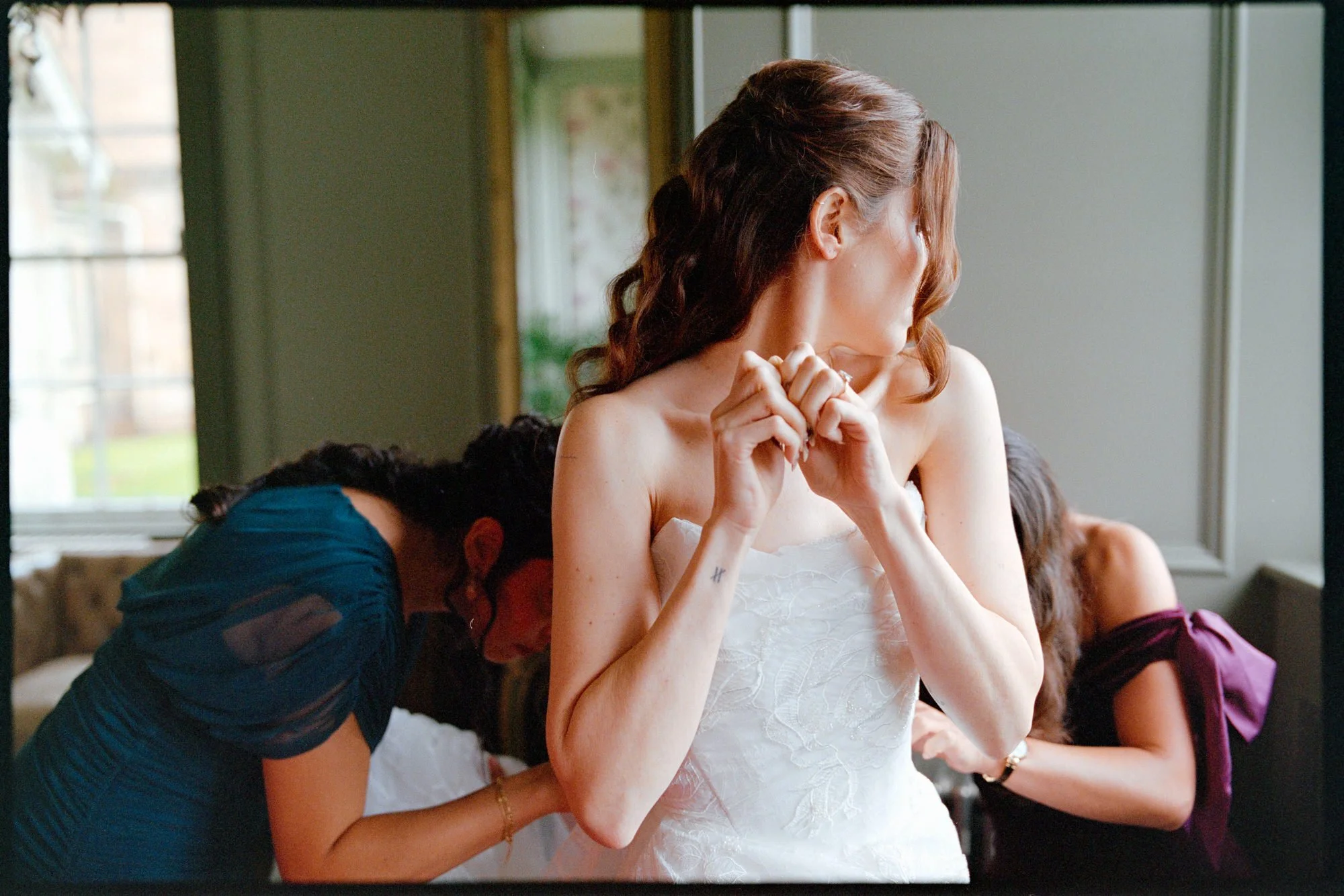 A bride wearing a white wedding dress stands with her hands clasped near her face while two women assist her with her dress, in a room with a window and natural light.