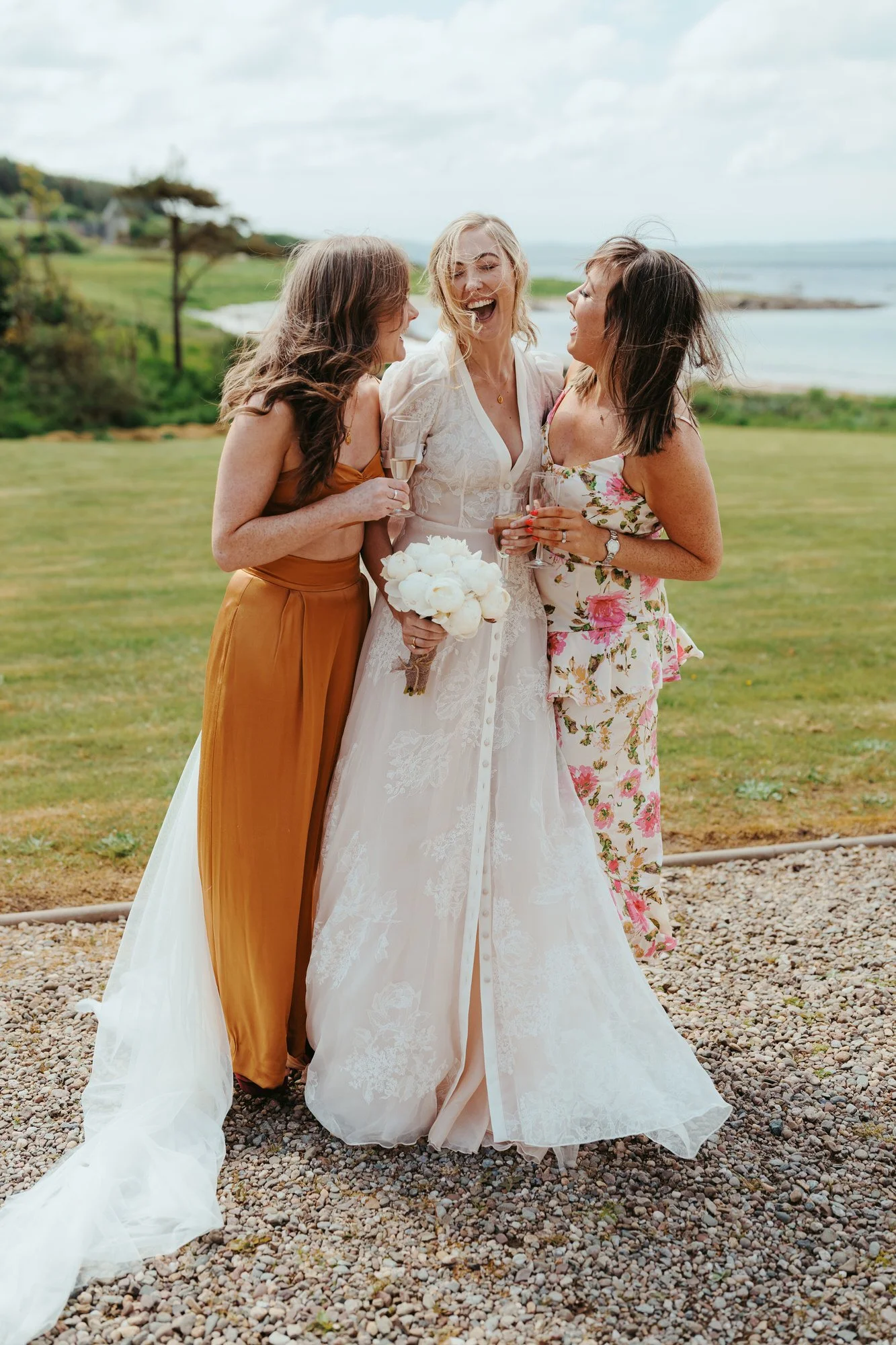 Three women, including a bride in a white wedding dress holding a bouquet, celebrating outdoors with two friends, all smiling and holding glasses of champagne near a waterfront backdrop. Photographed by Steven William Weir, Edinburgh Scotland.