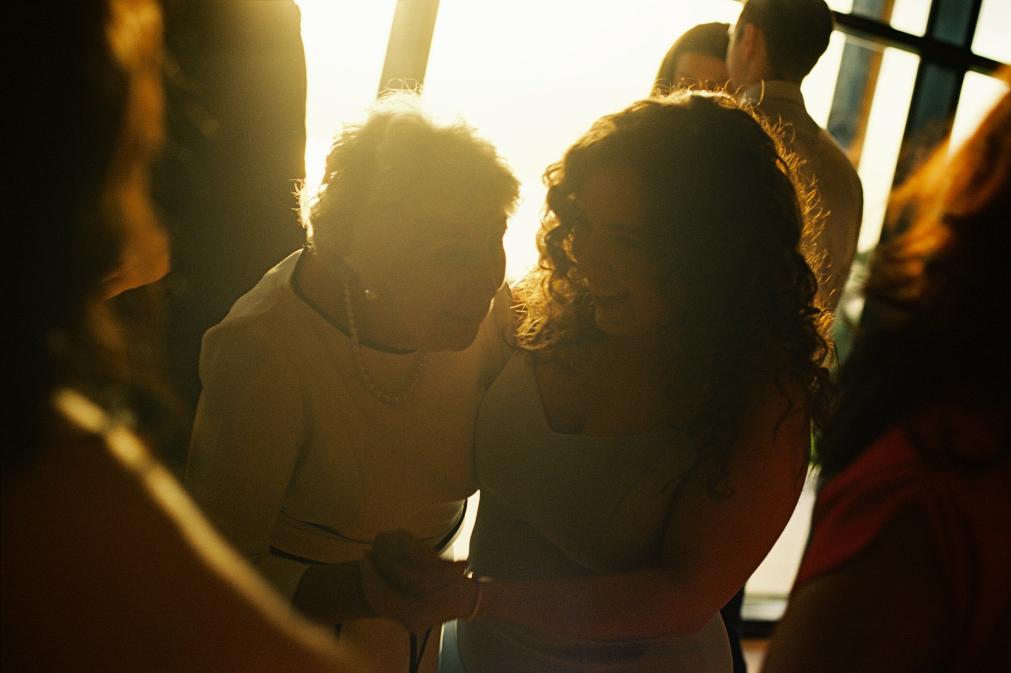Two women smiling and holding hands in a warm, backlit setting with other people in the background. Photographed by Steven William Weir, Edinburgh Scotland.